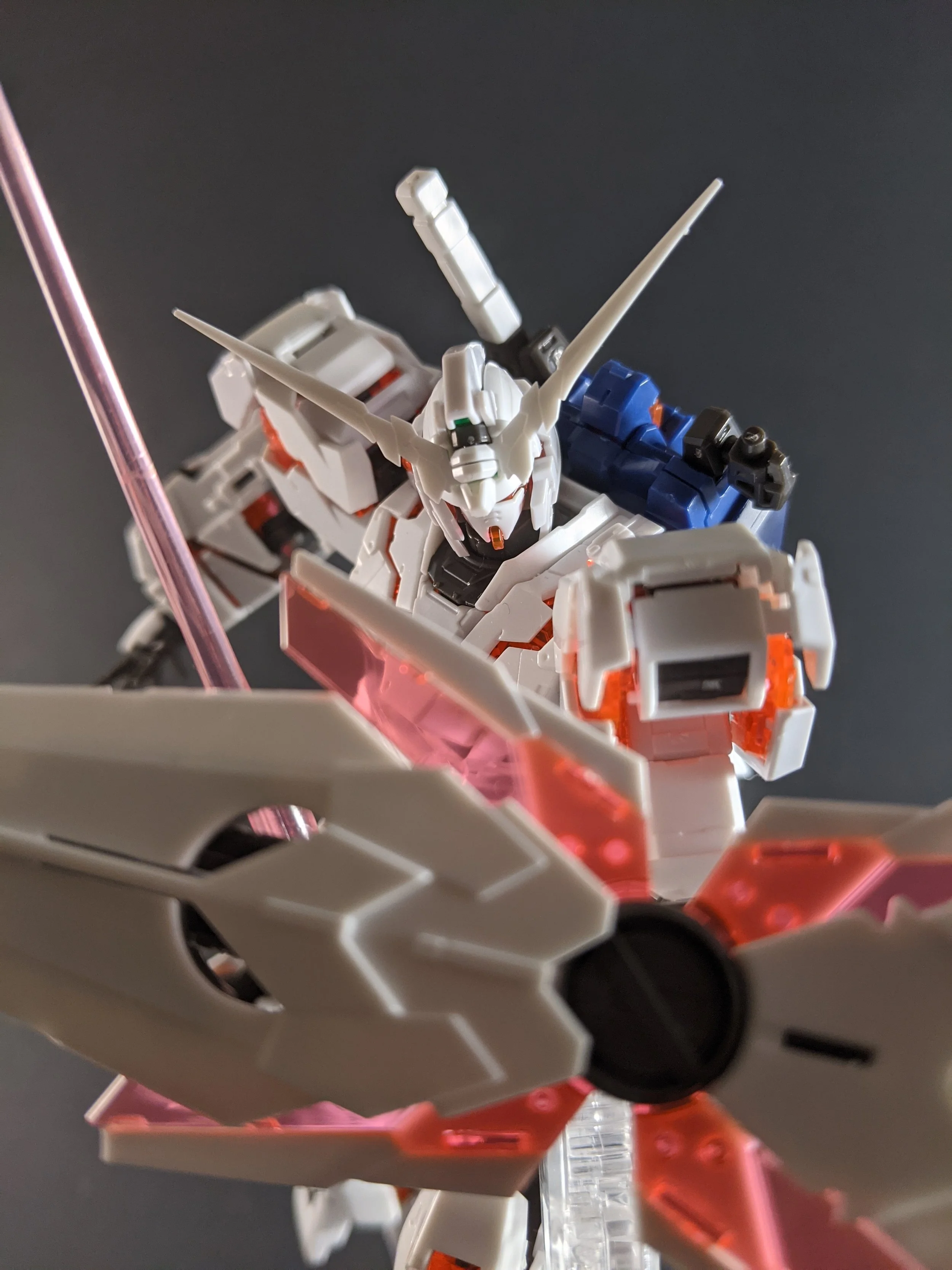 A detailed model of a white and blue robotic gundam with orange accents, wielding a large white sword with black and pink details, against a dark background.