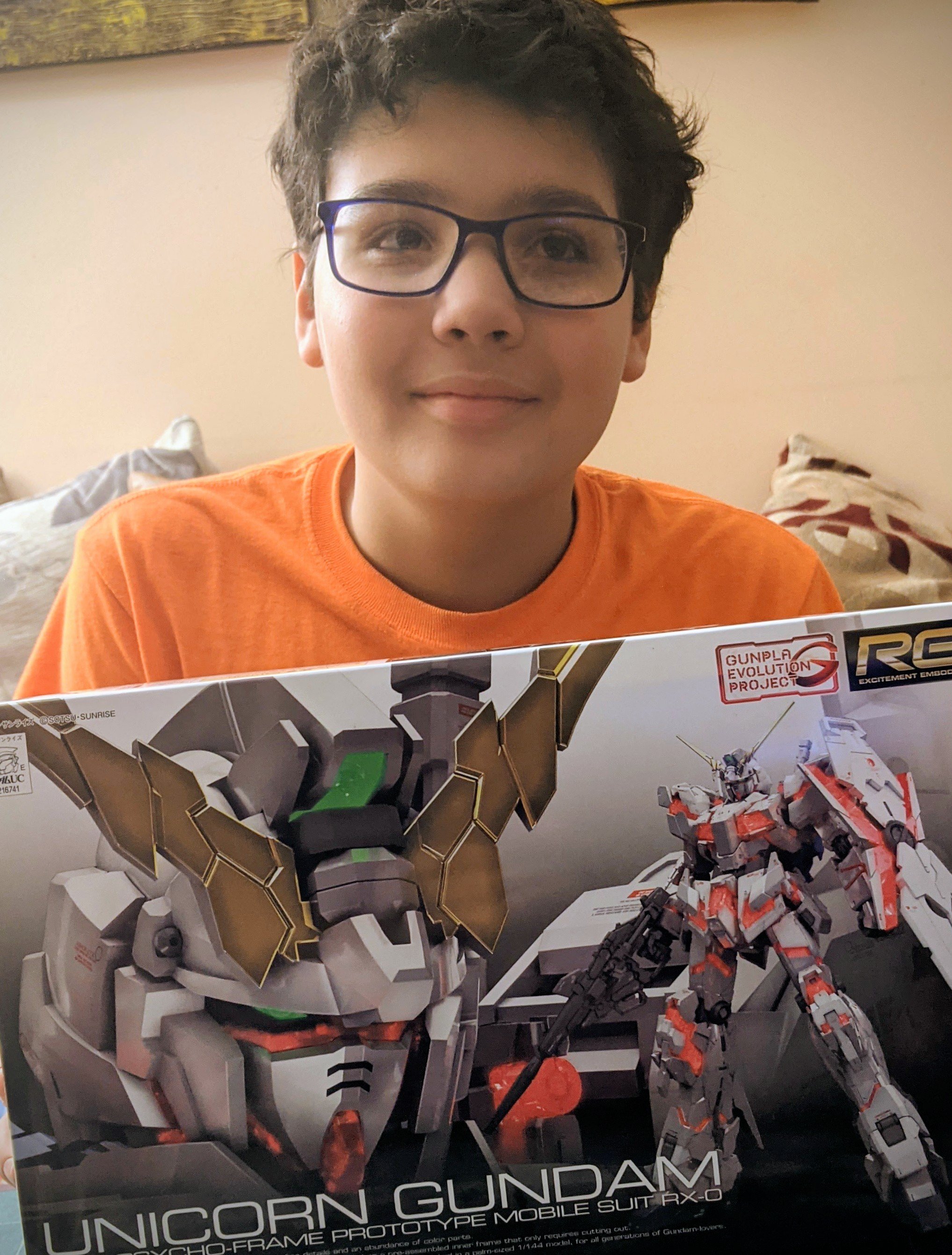 A young person with glasses holding a box of the Unicorn Gundam model kit. The box features artwork of a white and orange mobile suit with weaponry, labeled 'Unicorn Gundam'. The person is sitting on a couch with pillows in the background.