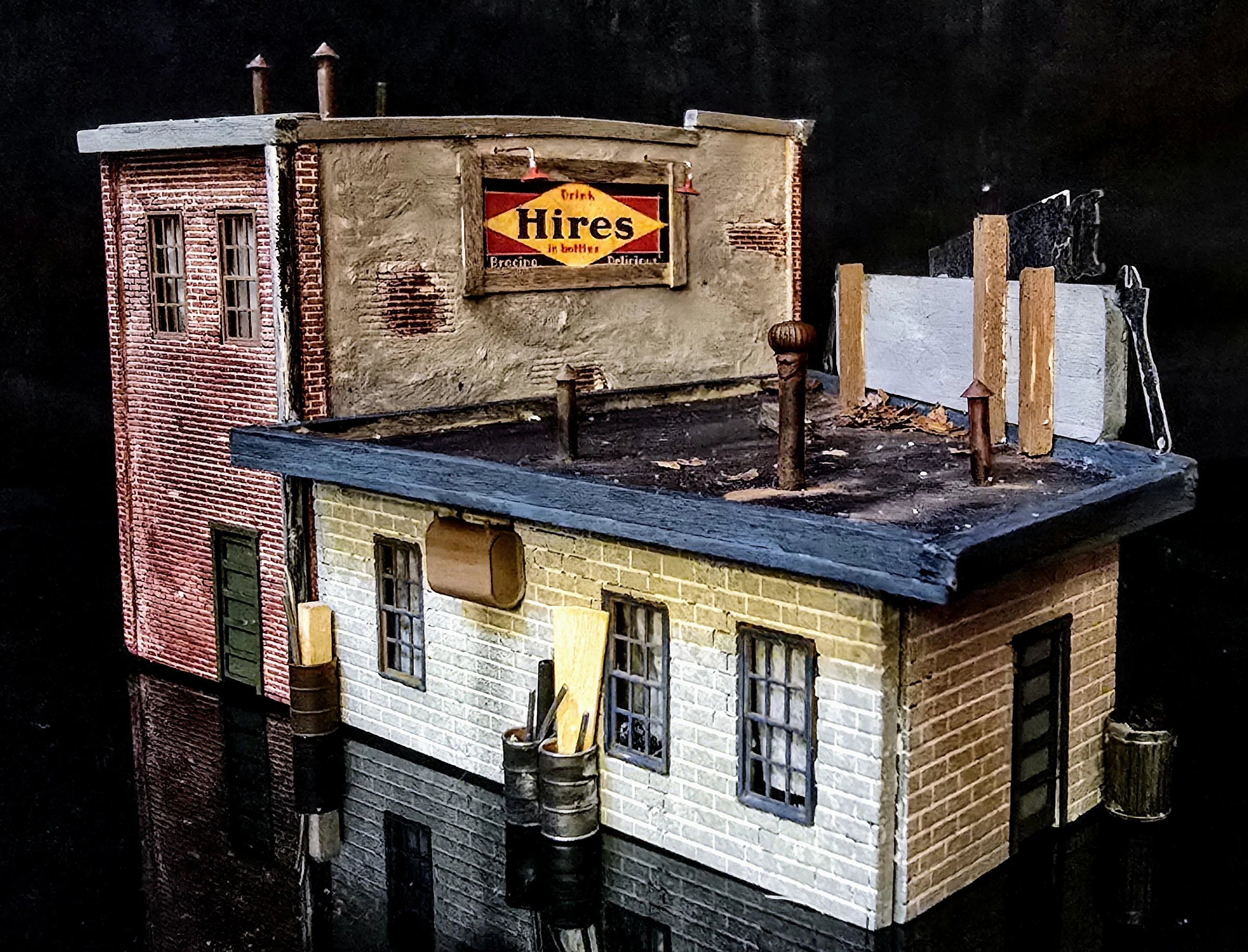 Miniature model of a two-story building with brick and stucco exterior, multiple windows, and a rooftop sign advertising "Hires" drink. There are wooden posts and structures on the roof, with some tools and debris nearby.