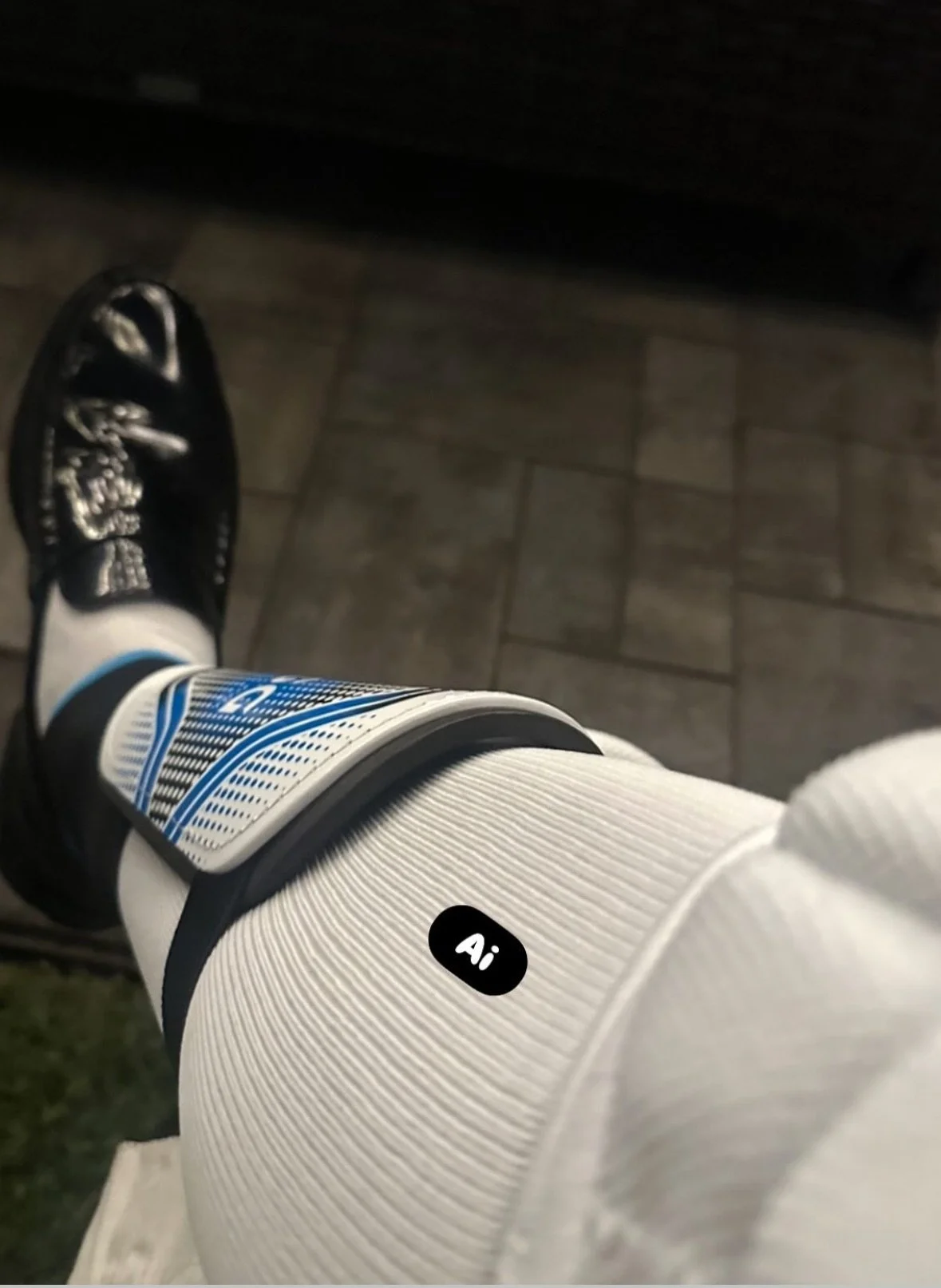Close-up of a person's leg wearing a white sports sock with a black label that says 'Ai', black athletic shoe, and a knee brace with blue and white designs, standing on a tiled floor.