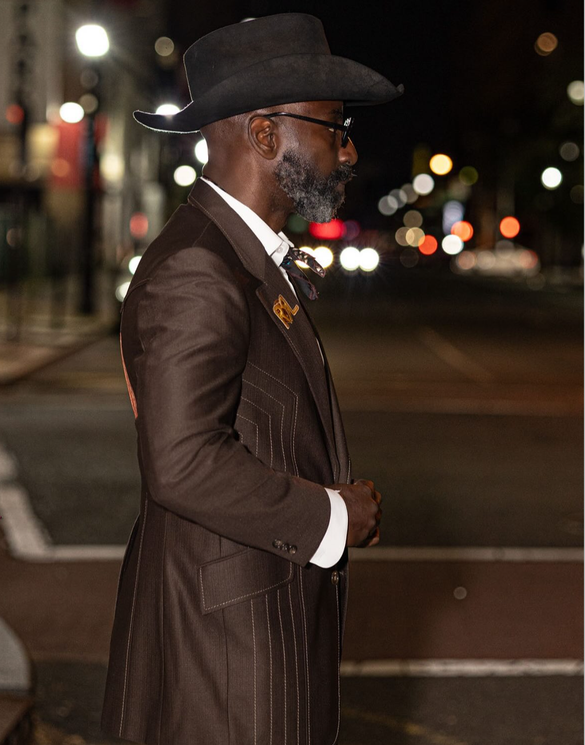 A man in a brown pinstripe suit, black hat, glasses, and with a beard is standing on a city street at night. The background is blurred with city lights.