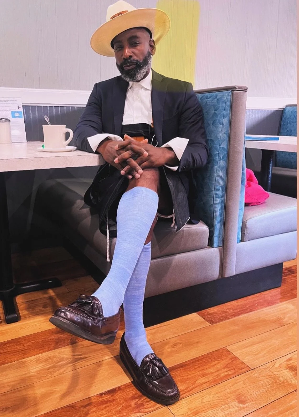 A man sitting in a restaurant booth, wearing a suit, white shirt, a wide-brimmed hat, light blue socks, and dress shoes, with his hands clasped, and one leg resting on the seat. There is a white mug and a napkin dispenser on the table.
