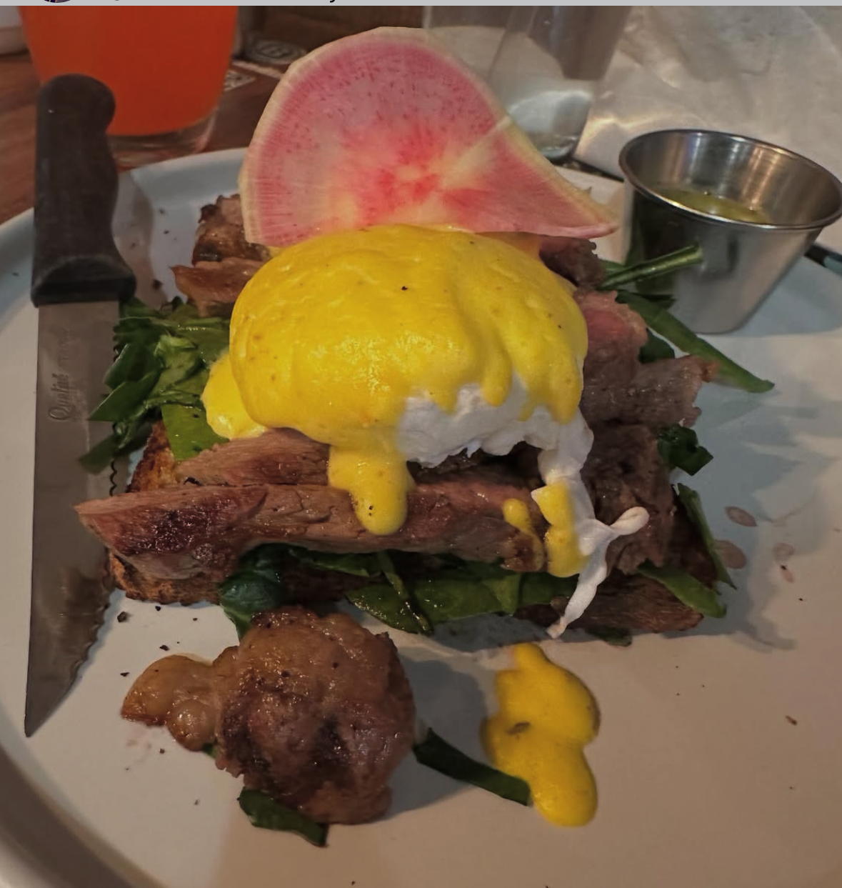 Steak and eggs breakfast with a poached egg topped with hollandaise, sliced watermelon radish, green vegetables, and a small cup of sauce on a plate.