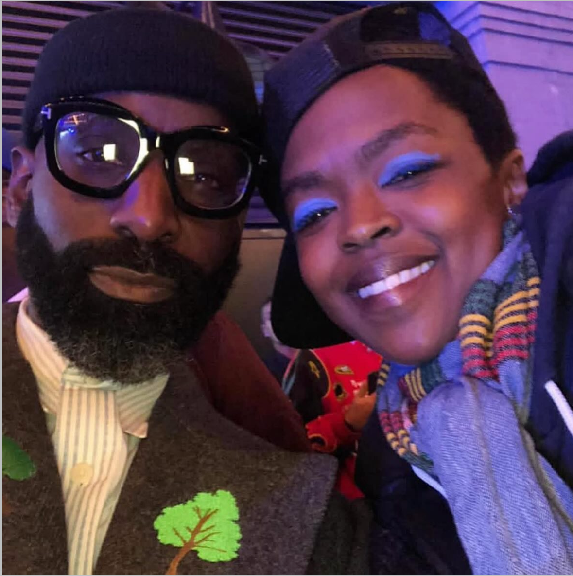 A man with glasses, a beard, and a beanie, and a woman with bright blue eyeshadow and a colorful scarf, taking a close-up selfie together in a dimly lit setting.