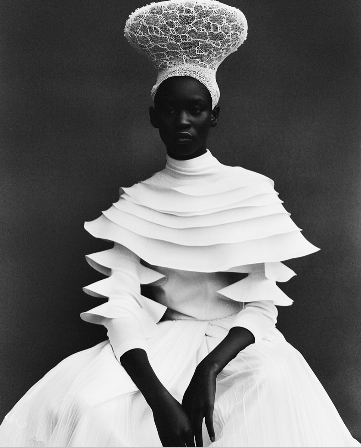 Black-and-white portrait of a woman with a large, intricate headpiece, wearing a layered, sculptural white dress with ruffled sleeves and pleated skirt, seated against a dark background.