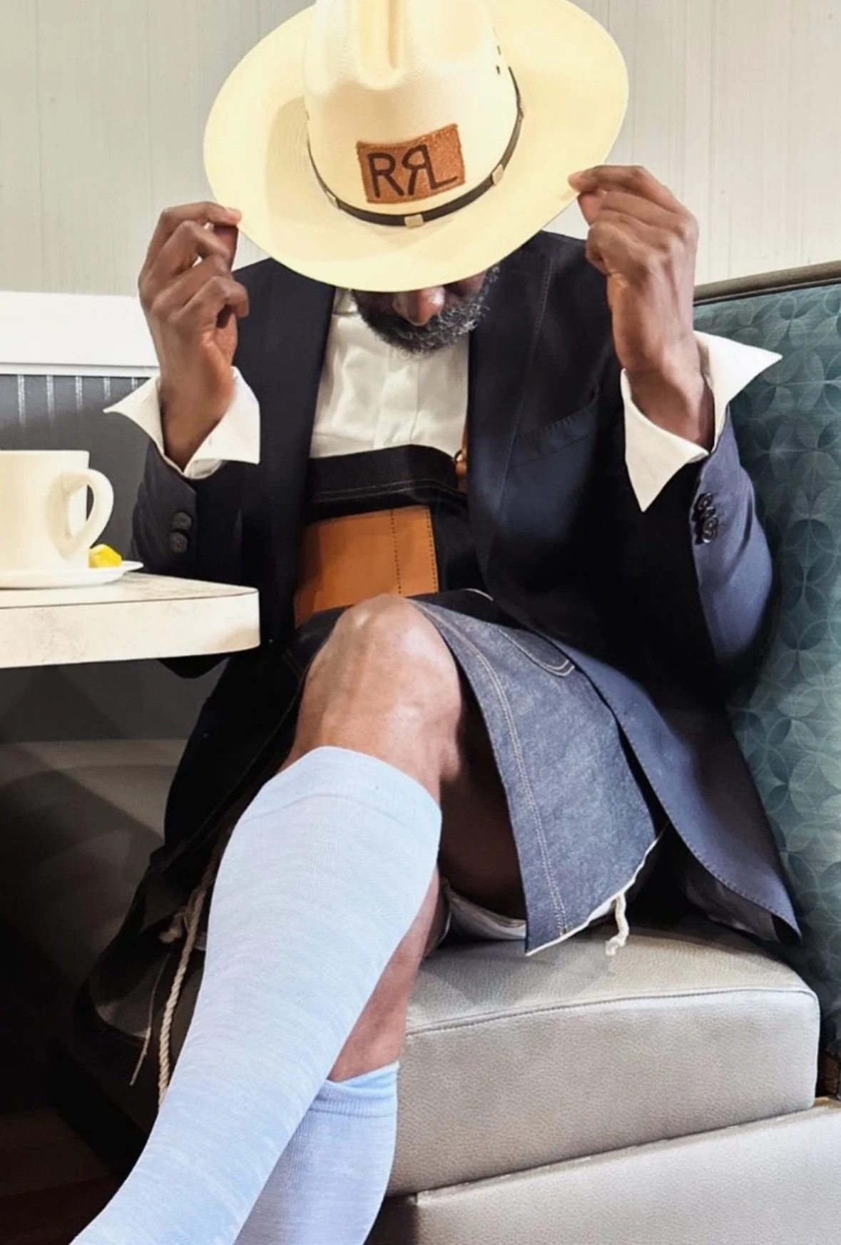 A man sitting in a booth at a restaurant, wearing a suit jacket, shorts, and a wizard hat. He is adjusting his hat and has a bandage on his knee, with a white sock pulled up to his knee.