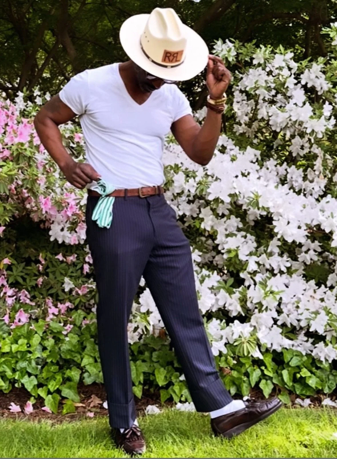 A man dressed in navy pinstriped pants, a white t-shirt, and brown shoes, standing outdoors next to blooming azalea bushes. He is holding a striped tie in one hand and adjusting a white cowboy hat with the other.