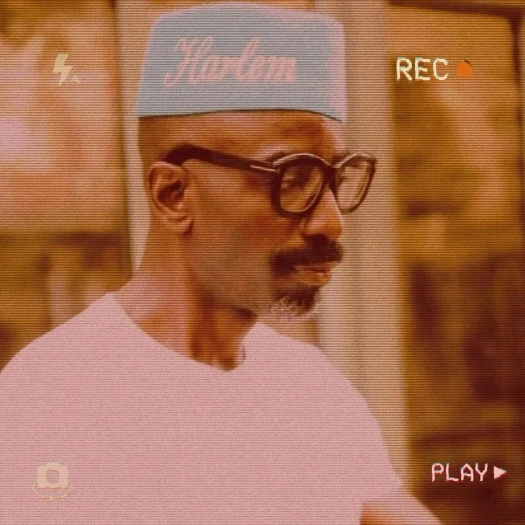 A man with glasses and a mustache wearing a light blue cap with pink text, and a light pink shirt, seen through a vintage video recorder screen.