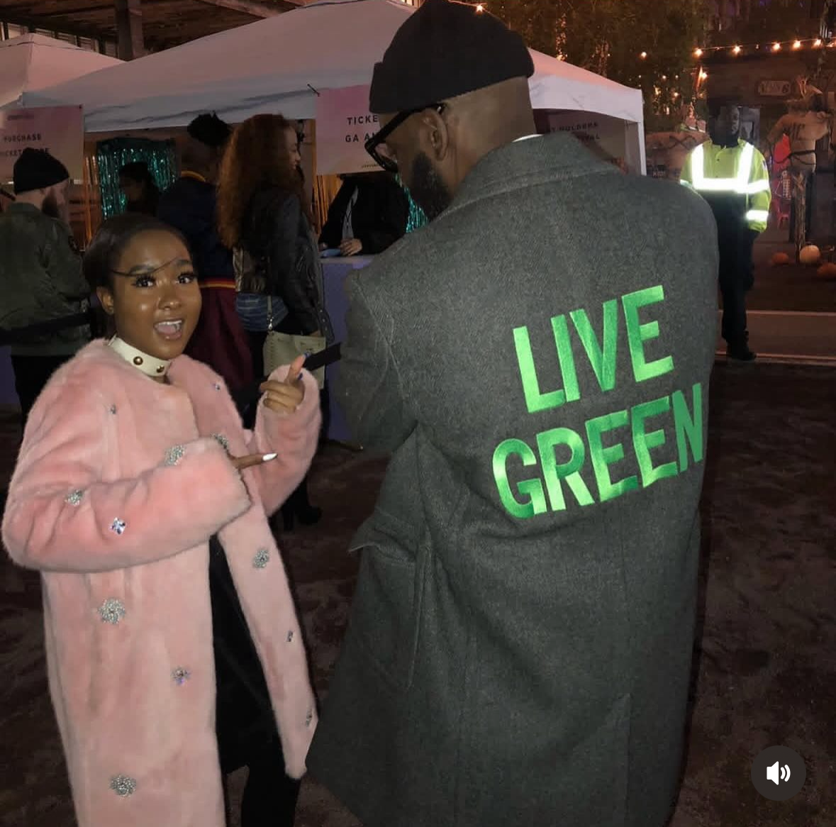 A woman in a pink coat with embellished details is speaking to a man wearing a grey blazer with the words 'LIVE GREEN' in bright green on the back during an outdoor event at night. There are tents and other people in the background.