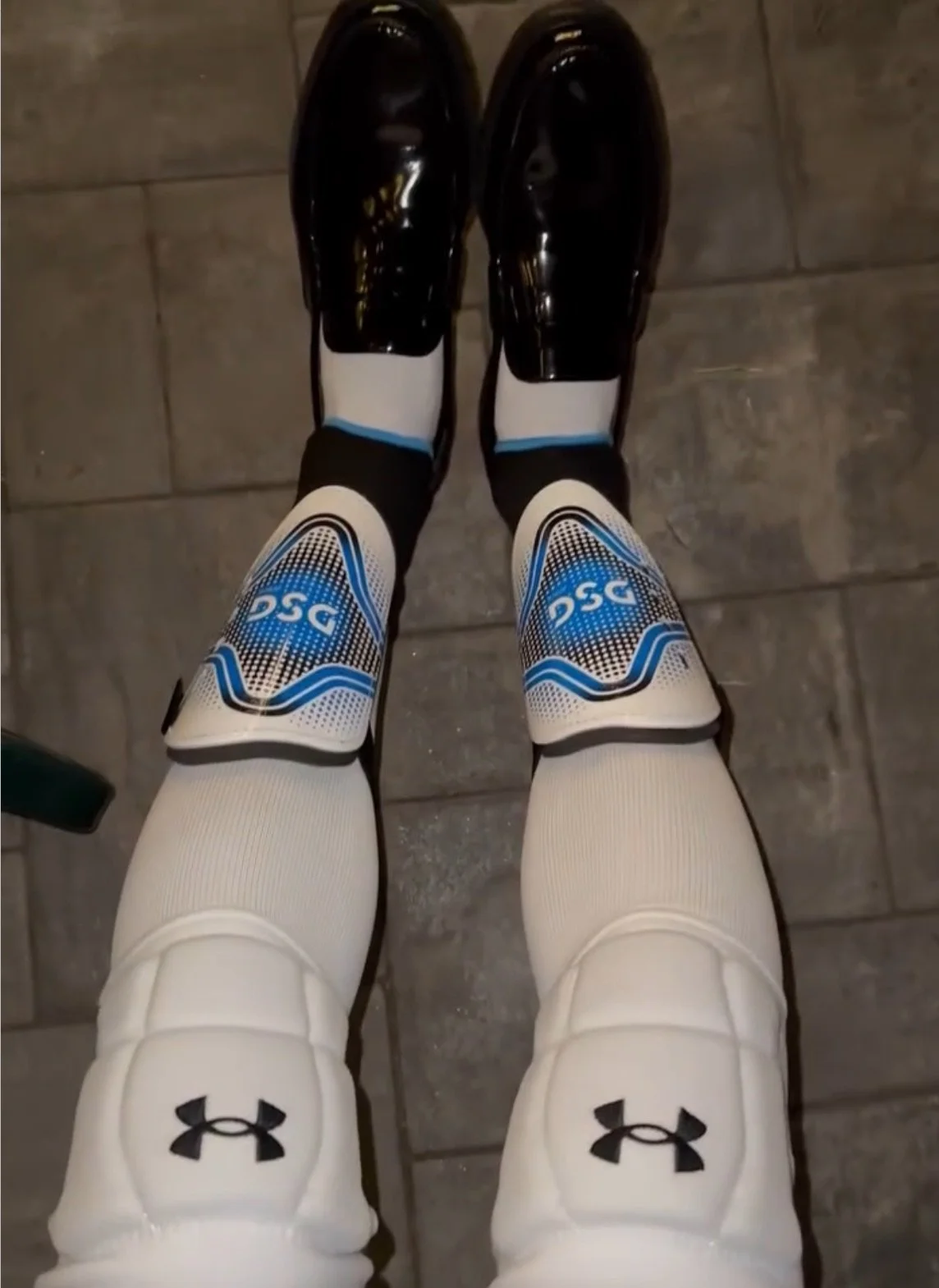 A person wearing white Under Armour compression tights, black and blue DSC cycling shoes, and black shin guards, standing on a tiled floor.