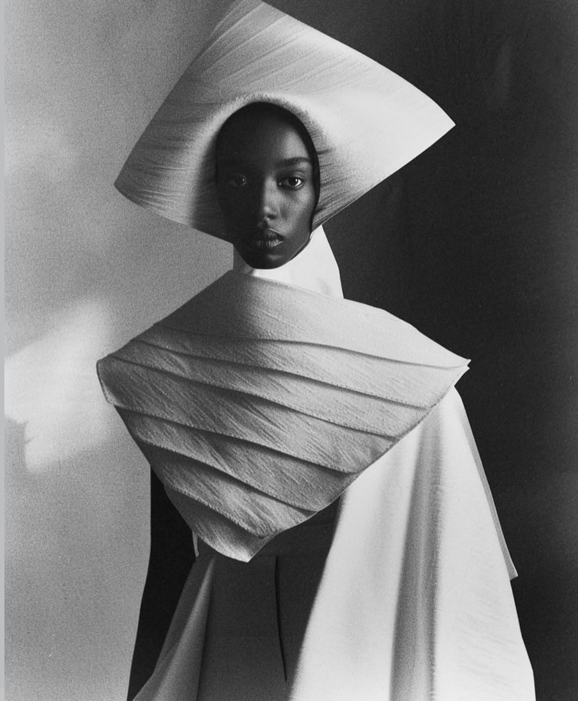 A black and white photograph of a woman with a stylized geometric headpiece and dress, creating an abstract, artistic look.