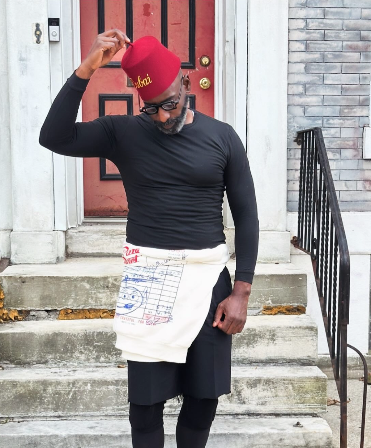 A man standing on front steps in front of a red door and gray brick wall, wearing a red chef's hat and apron, black long-sleeve shirt, black shorts, and glasses, adjusting his hat.