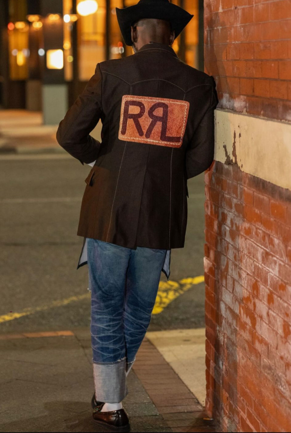 Man wearing a black cowboy hat, black jacket with a patch on the back with red and dark purple letters 'RRL', standing next to a brick wall on a city street at night.