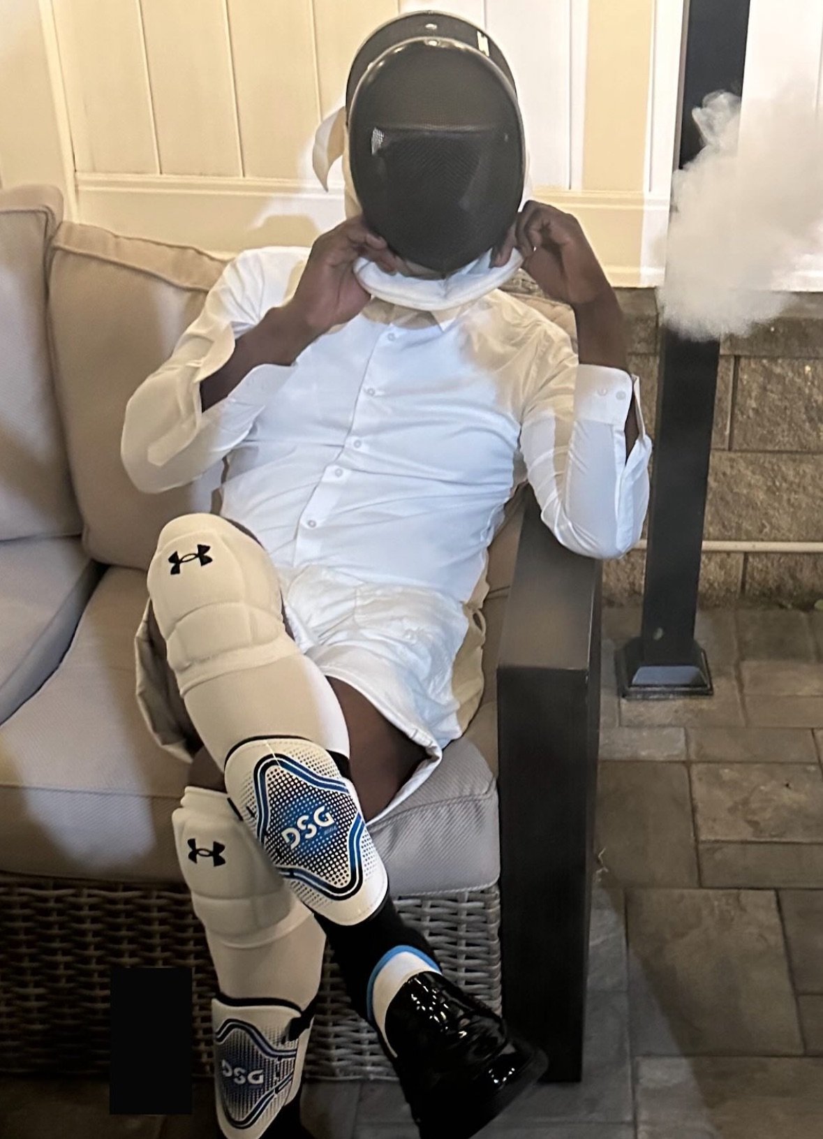 A person sitting on a couch, wearing a fencing mask, white long-sleeve shirt, athletic knee-high socks, and black shoes, with steam or vapor coming from the mask