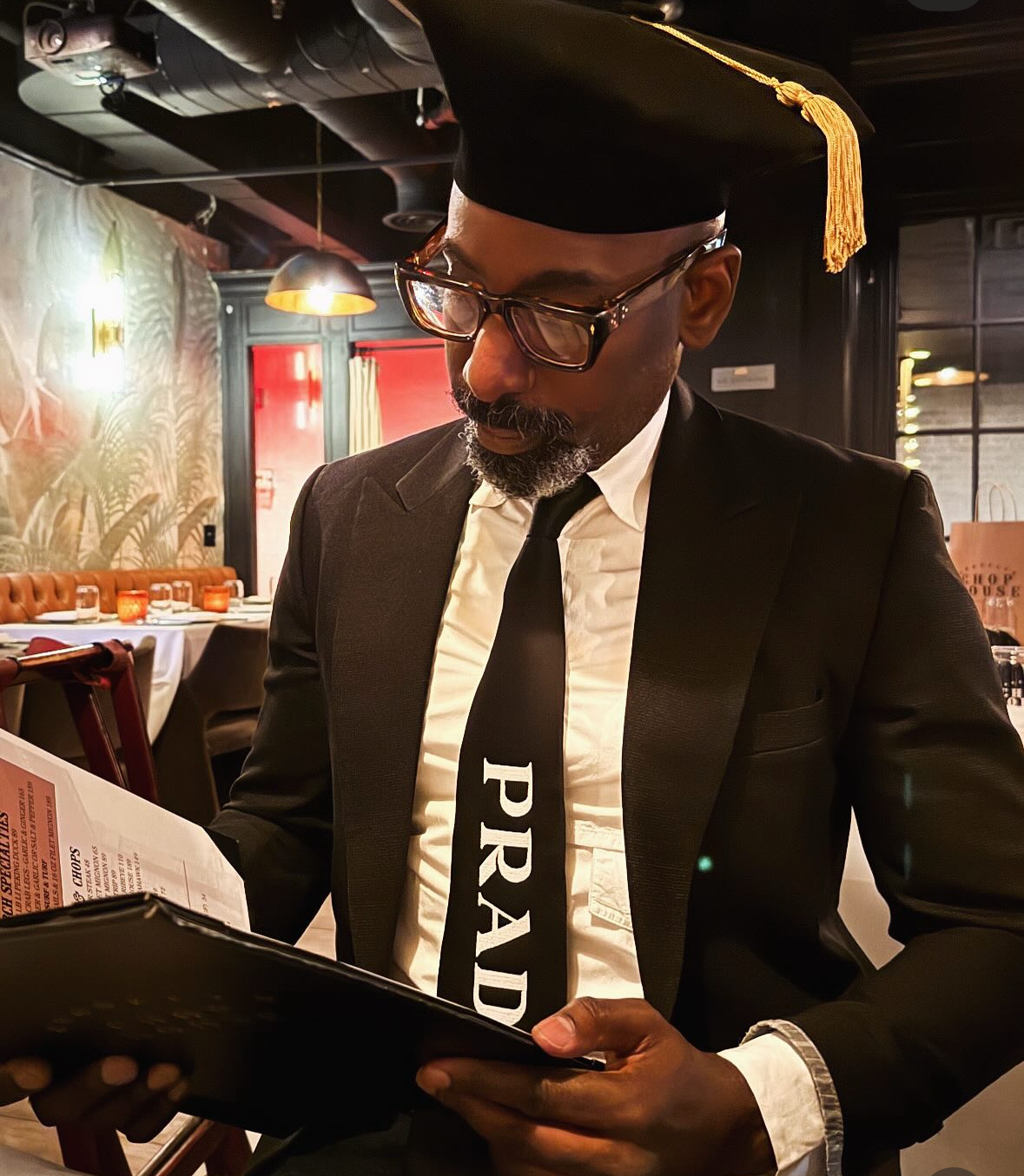 A man dressed in a suit and tie, wearing a graduation cap, is reading a menu in a restaurant.