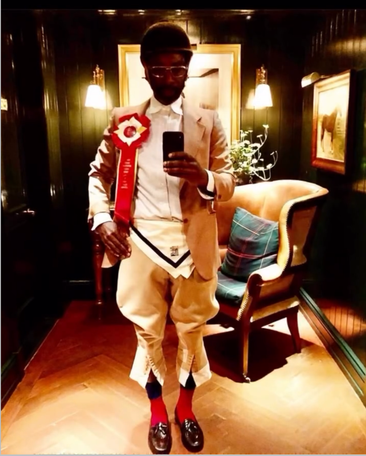 A man in a beige suit, white shirt, and dress shoes with red socks, standing in a warmly lit room with dark wood paneling, taking a mirror selfie. He has a ribbon badge on his chest and is holding a small bag in his left hand. There is a beige armcha