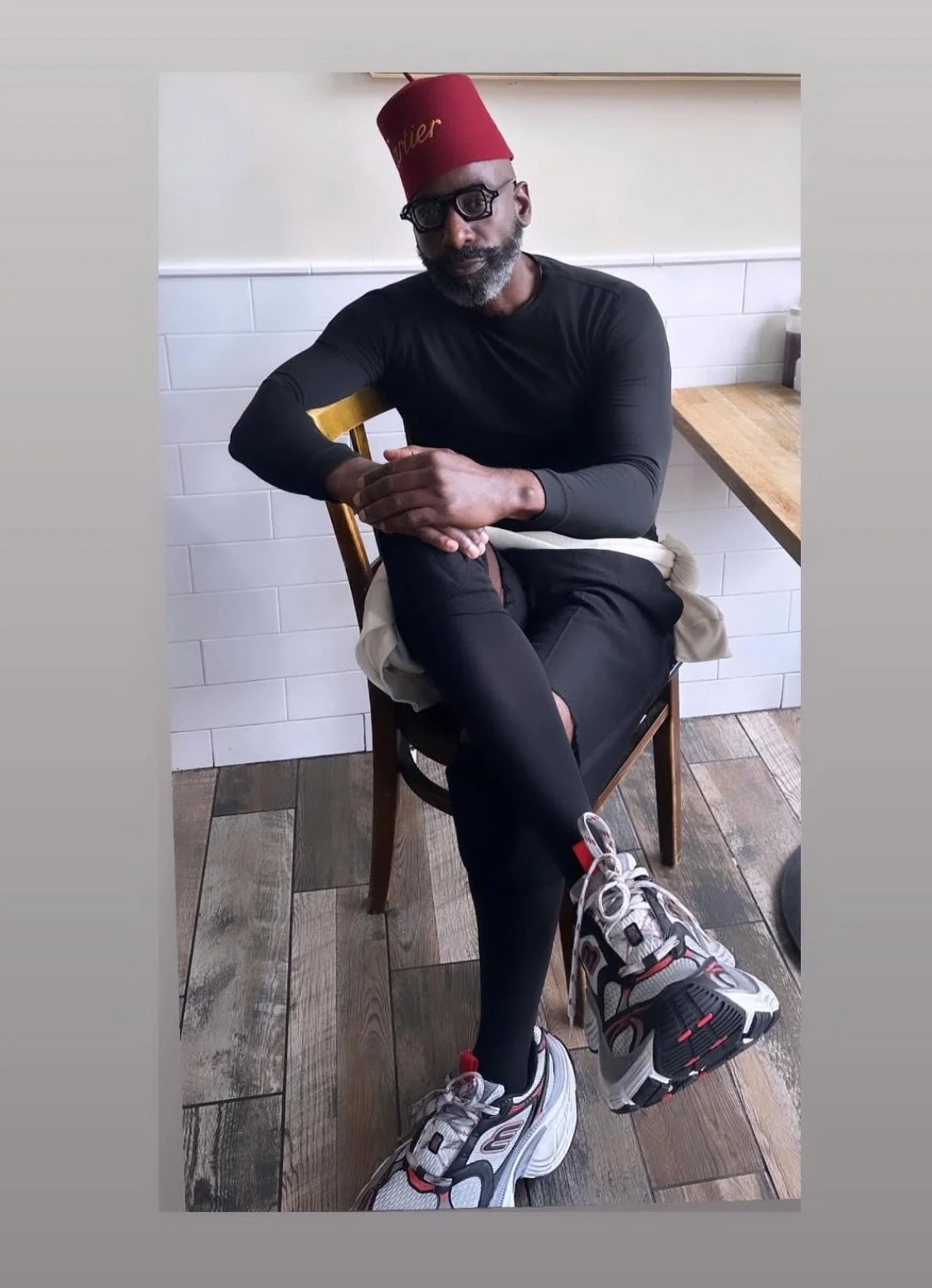 A man sitting at a restaurant table, wearing a squash hat, black sunglasses, a black long-sleeve shirt, black pants with a tear, and colorful sneakers, with one leg crossed over the other.