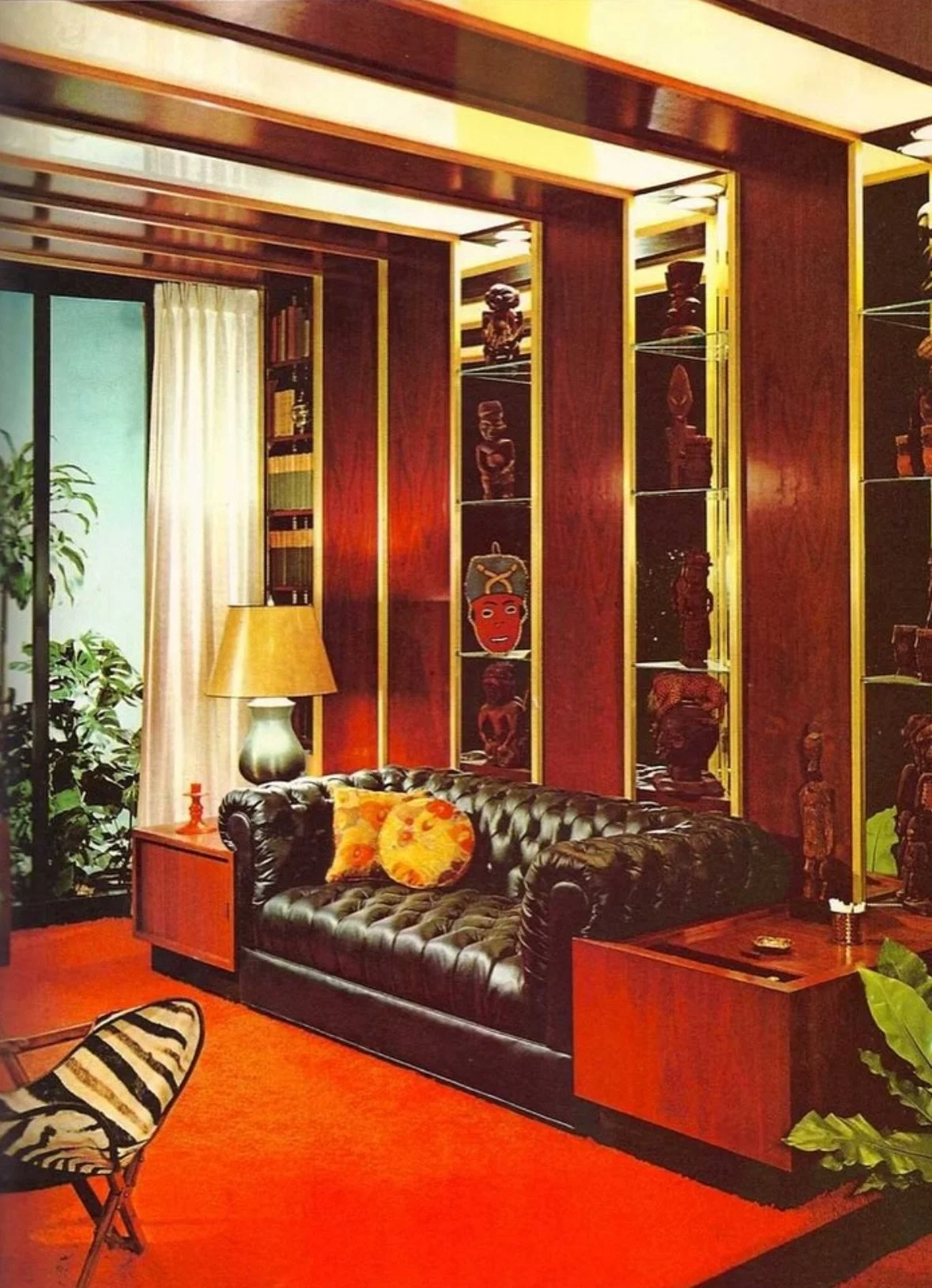 Living room with red carpet, black leather tufted sofa, orange pillows, wooden side tables, green plants, and a mantle with decorative statues and art pieces, illuminated by warm lighting.