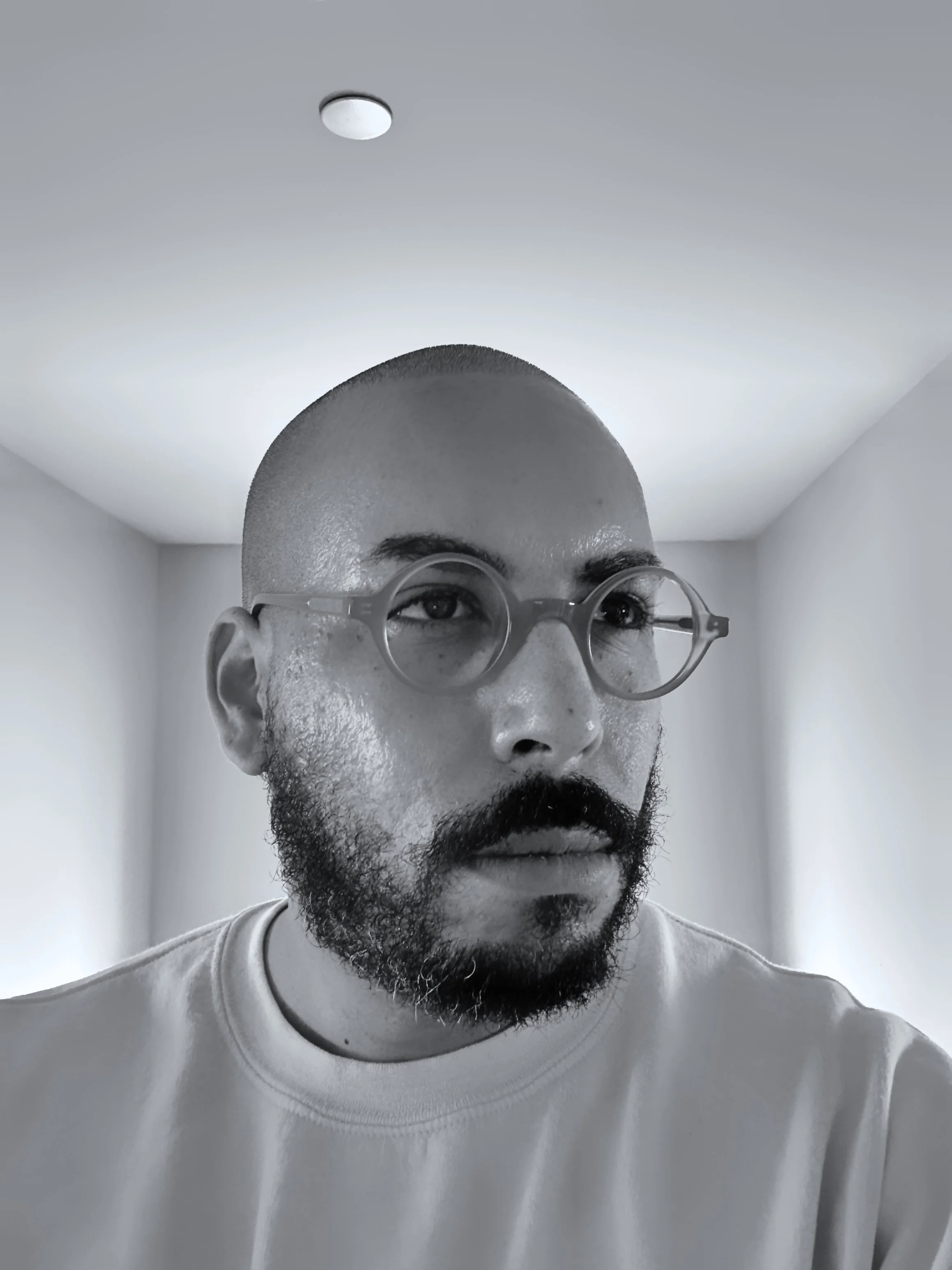 Supjamil with a shaved head, glasses, and a beard looking slightly to the side in a room with plain walls and a ceiling light