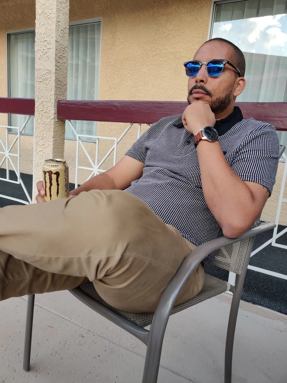 Supjamil sitting outside on a patio chair, wearing sunglasses, a checkered shirt, and khaki pants, holding a Monster energy drink can, with a beige building in the background.