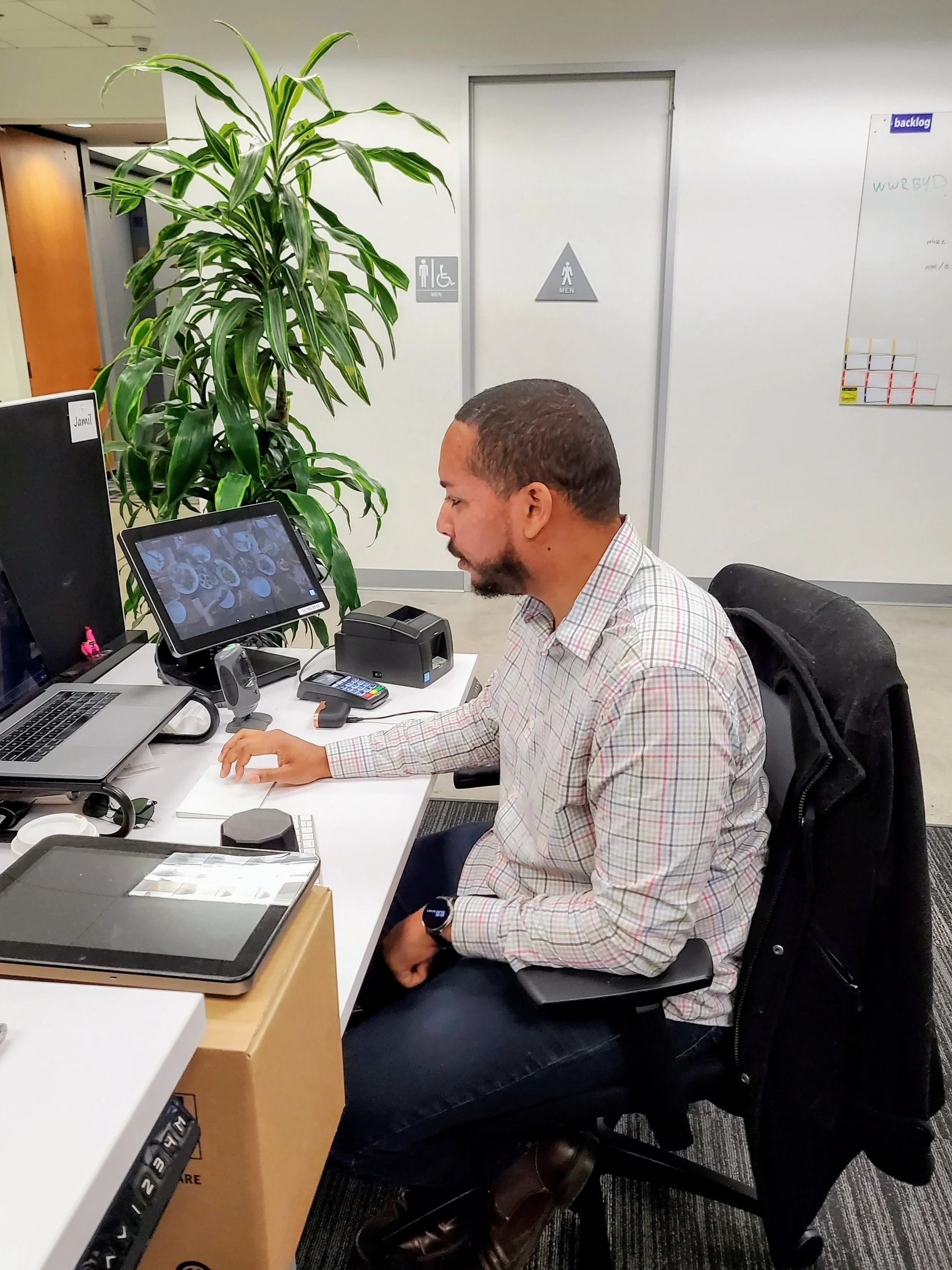 Supjamil sitting at a cluttered desk in an office, working on a computer with multiple monitors, surrounded by office equipment and a large green plant.