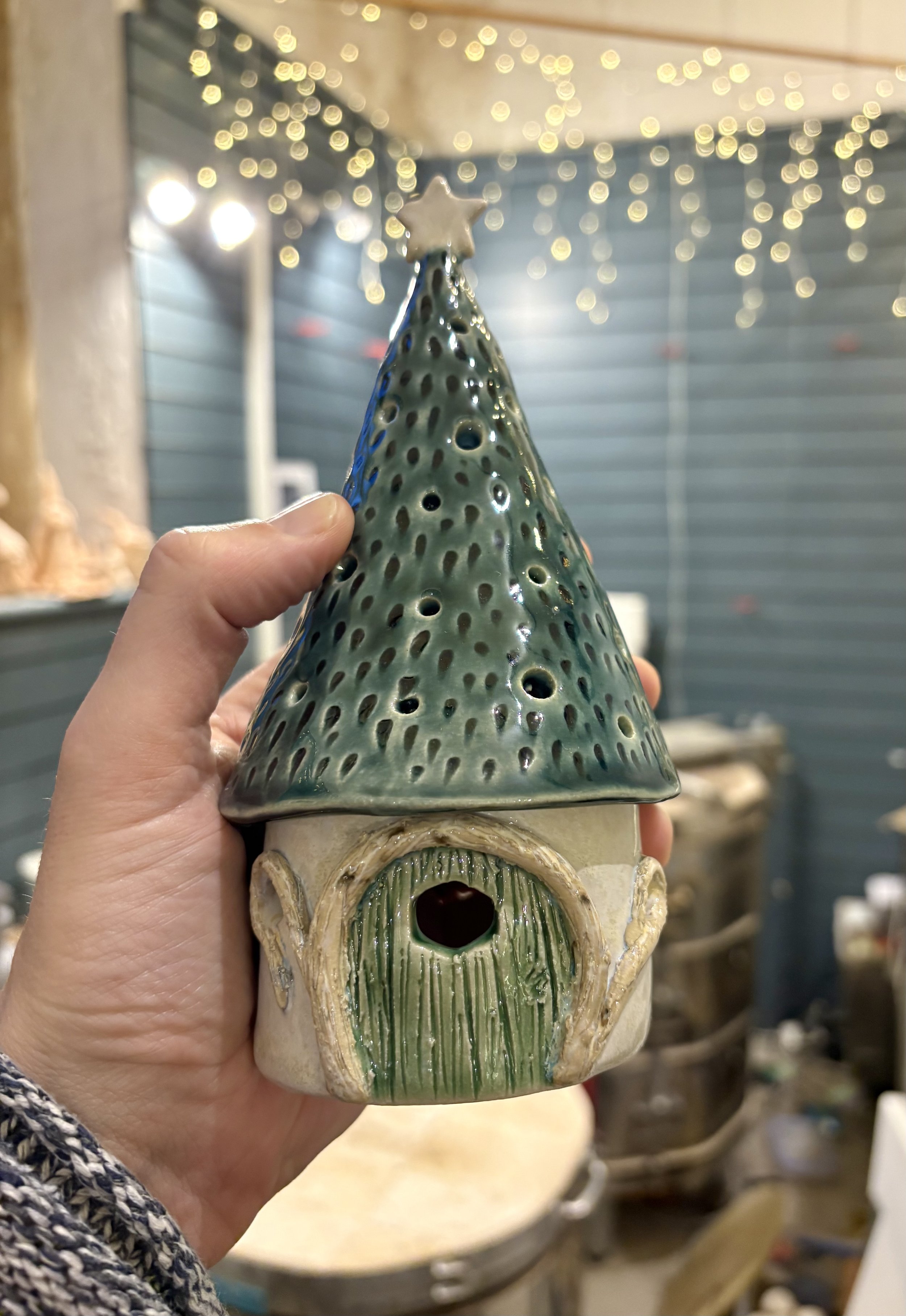 A ceramic fairy house with a green conical roof, intricate door, and star on top, held in hand against a blurred background with fairy lights.
