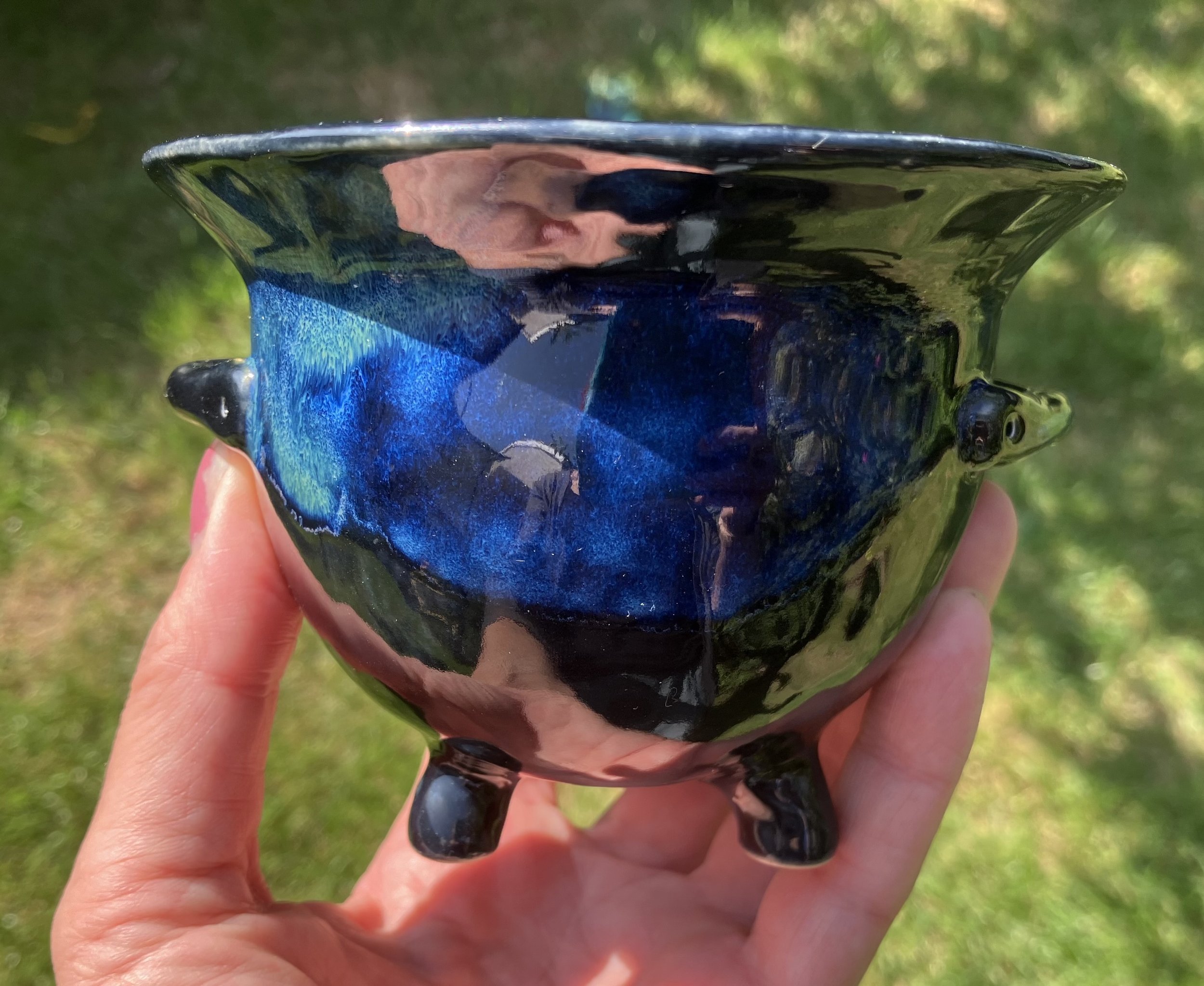 A small, black, glazed ceramic bowl with a glossy finish, held outdoors against a background of green grass, reflecting the sky and surrounding environment.