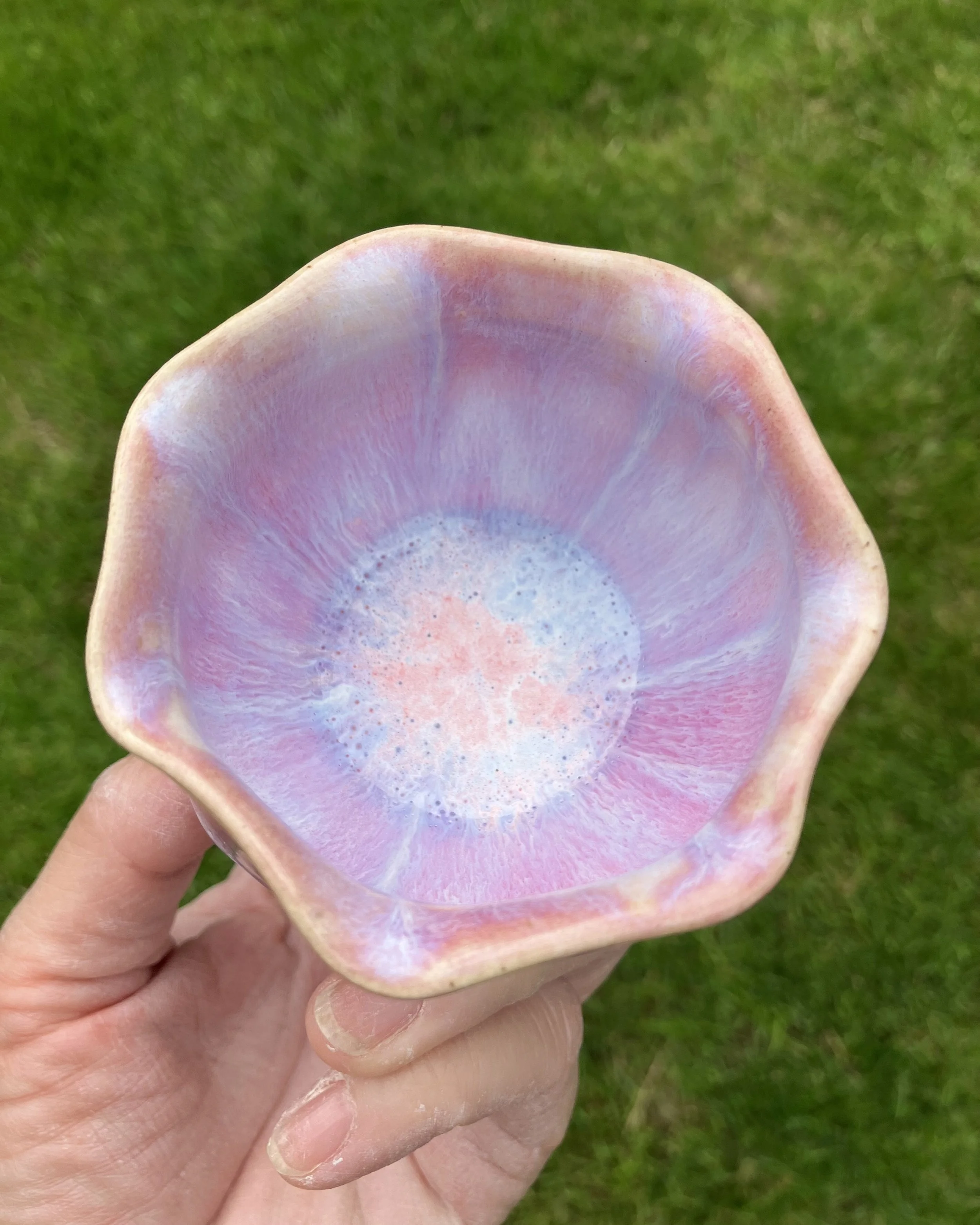 A hand holding a ceramic bowl with an iridescent pink, purple, and blue glaze, set against a green grassy background.