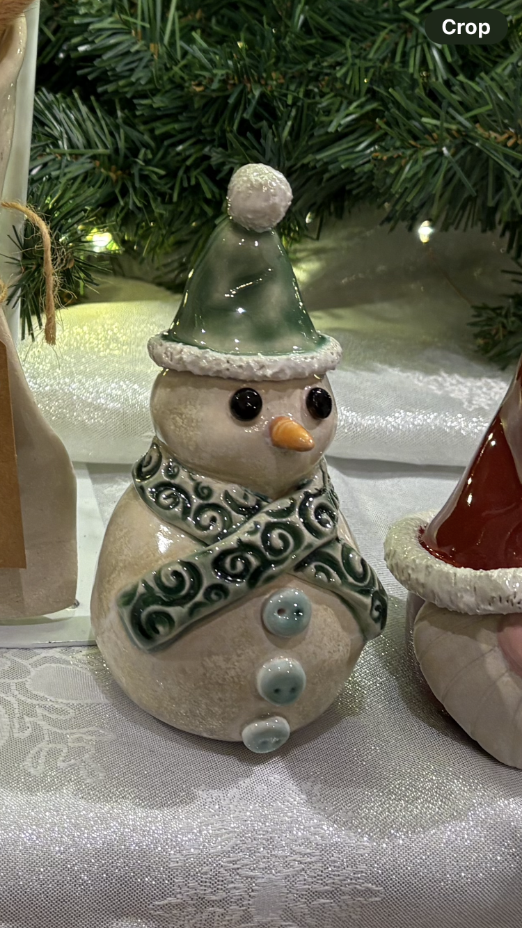 A snowman-shaped Christmas figurine wearing a green Santa hat with a white pom-pom, a patterned scarf, and three blue buttons. It has large black eyes and an orange nose.