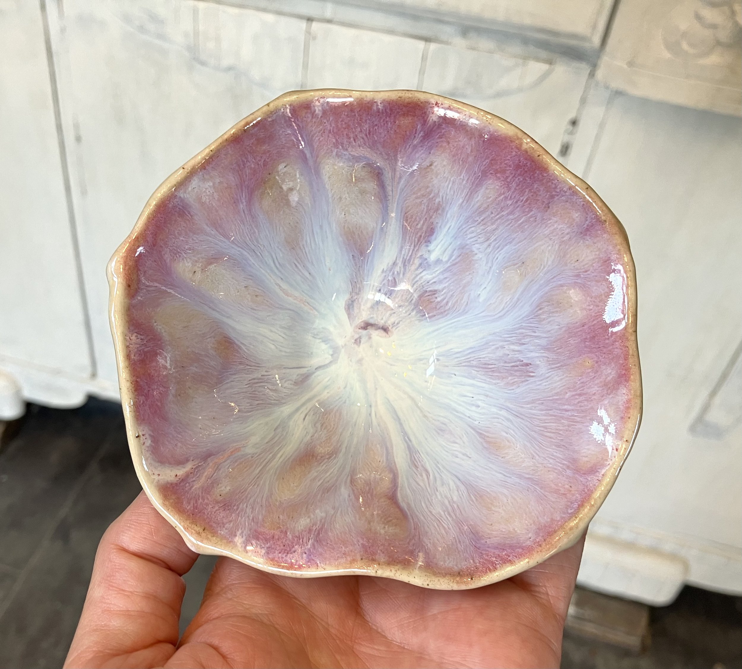 A ceramic bowl with an irregular, wavy edge displays a glaze mimicking the appearance of a lemon slice, featuring a pinkish outer rim and white, radiating streaks resembling fruit pulp.