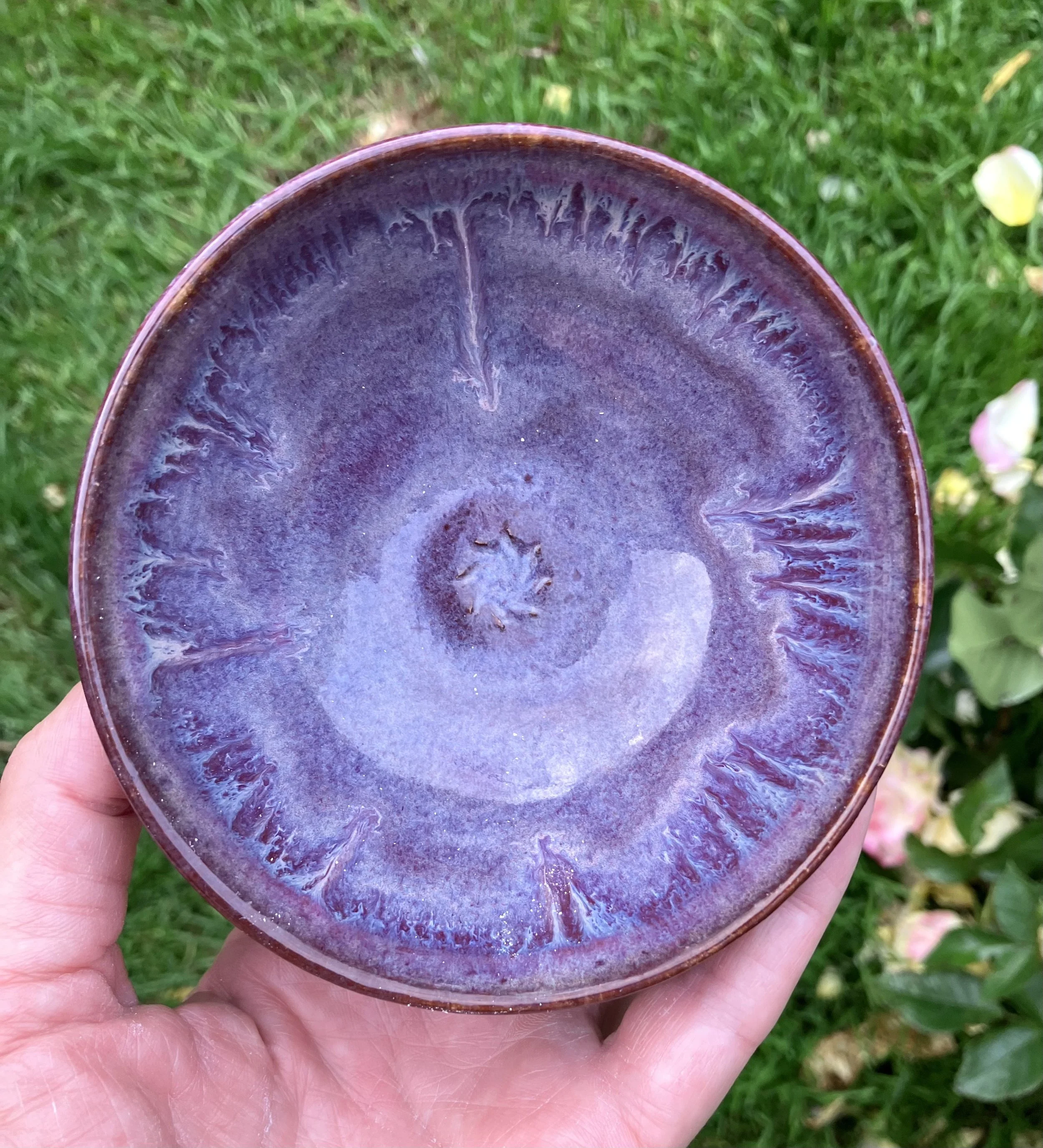 A person's hand holding a purple glazed ceramic bowl outdoors on green grass.
