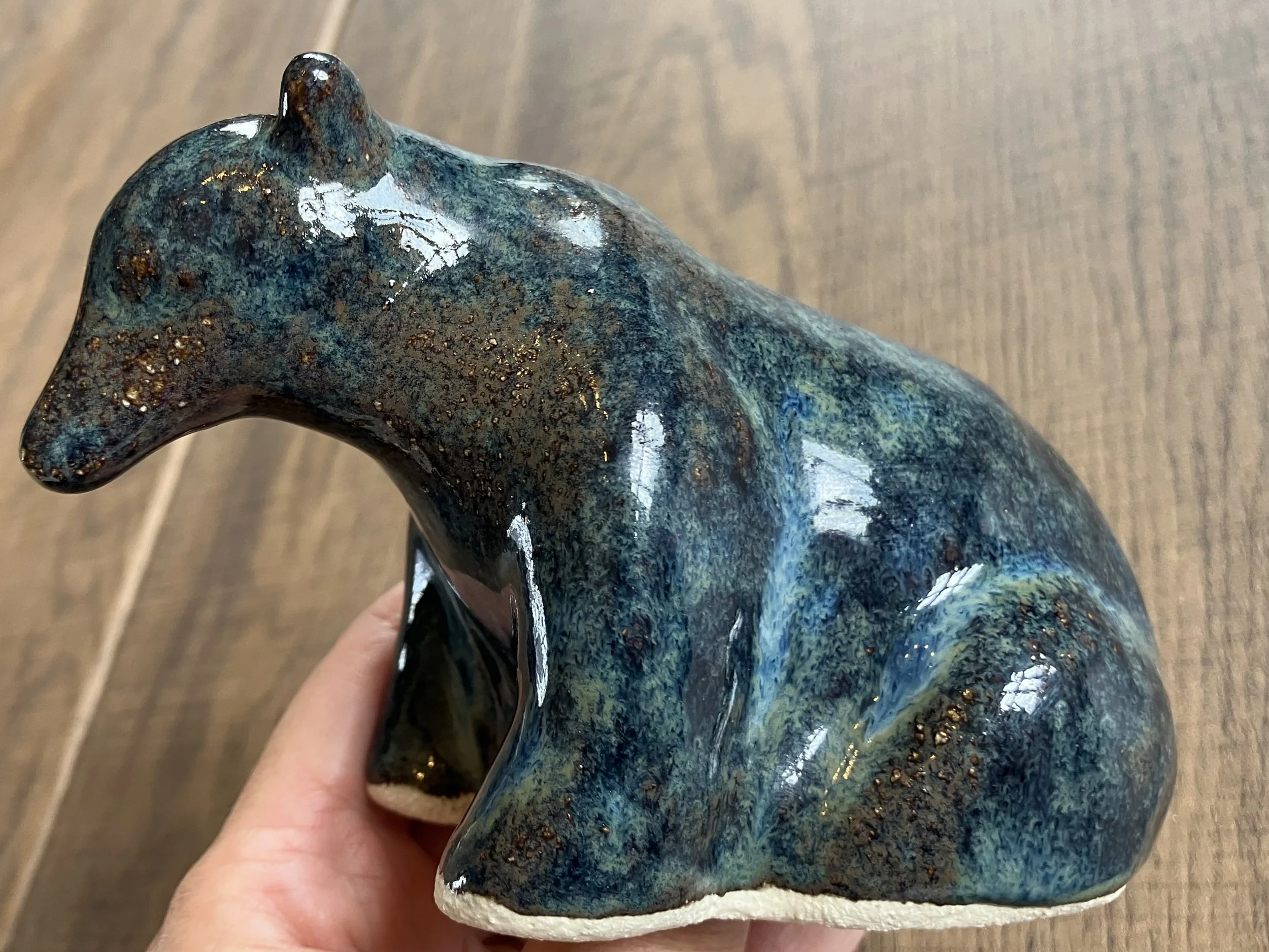 Ceramic sculpture of a stylized animal, possibly a bear, with a dark glossy glaze featuring blue, green, and brown tones, held by a person's hand over a wooden surface.