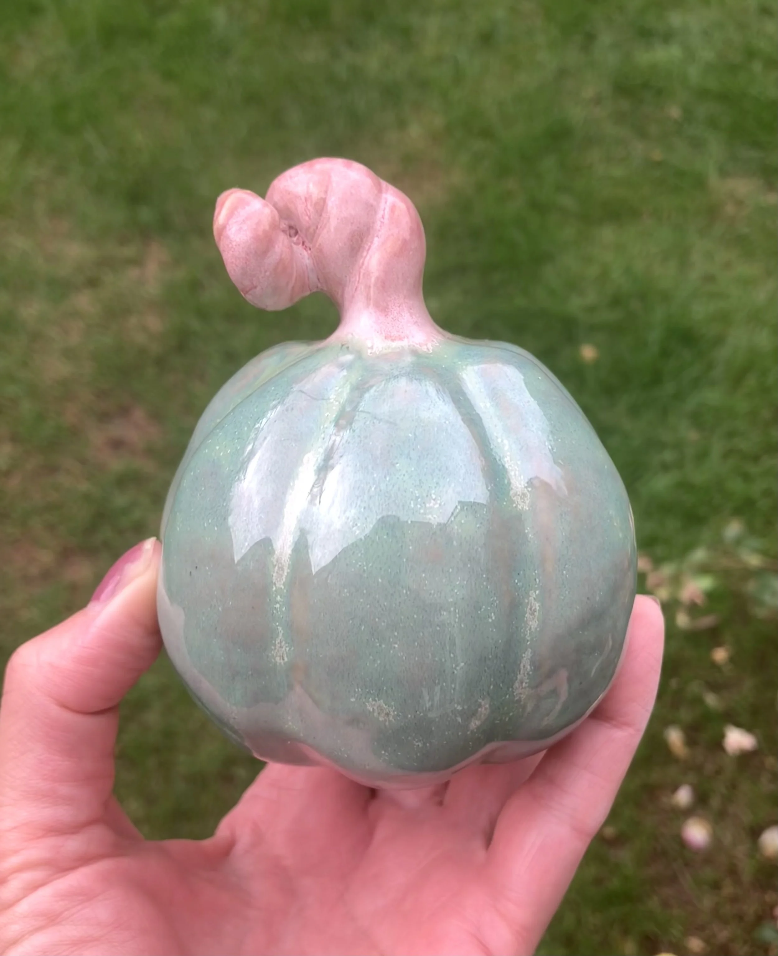 A ceramic pumpkin with a pinkish-green stem, held in a person's hand outdoors with grass in the background.