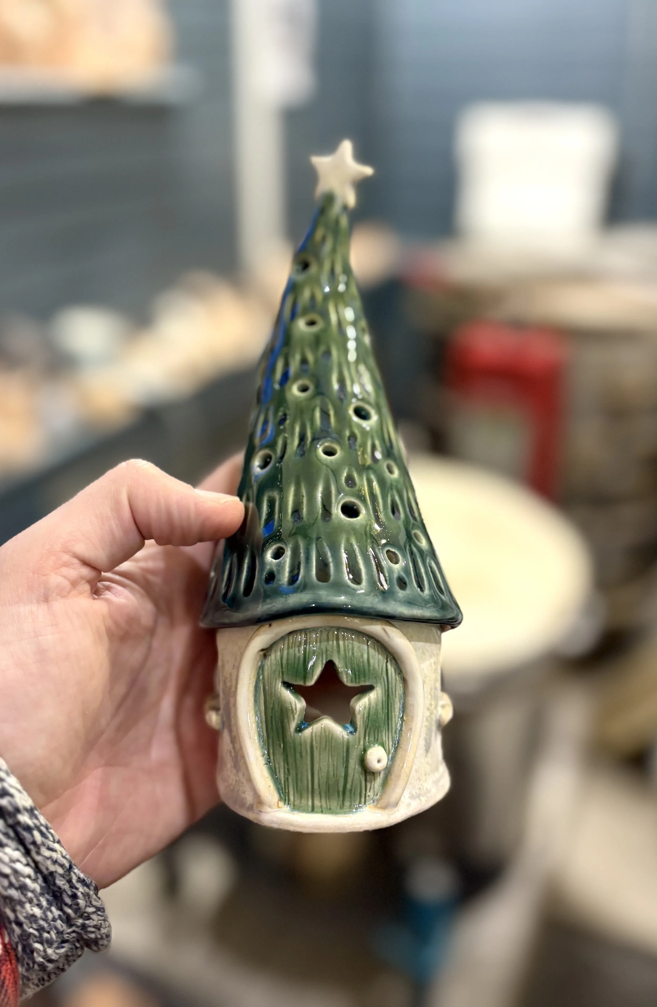A ceramic Christmas ornament shaped like a small house with a star-shaped cutout in the door and a conical, green, hole-filled roof topped with a star.