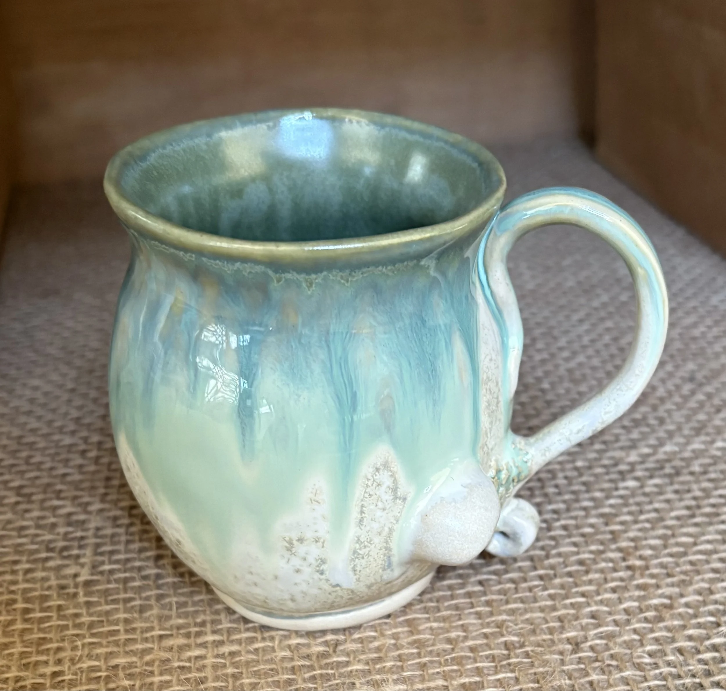 A ceramic mug with a glazed surface featuring blue, green, and cream drips, placed on a textured surface.