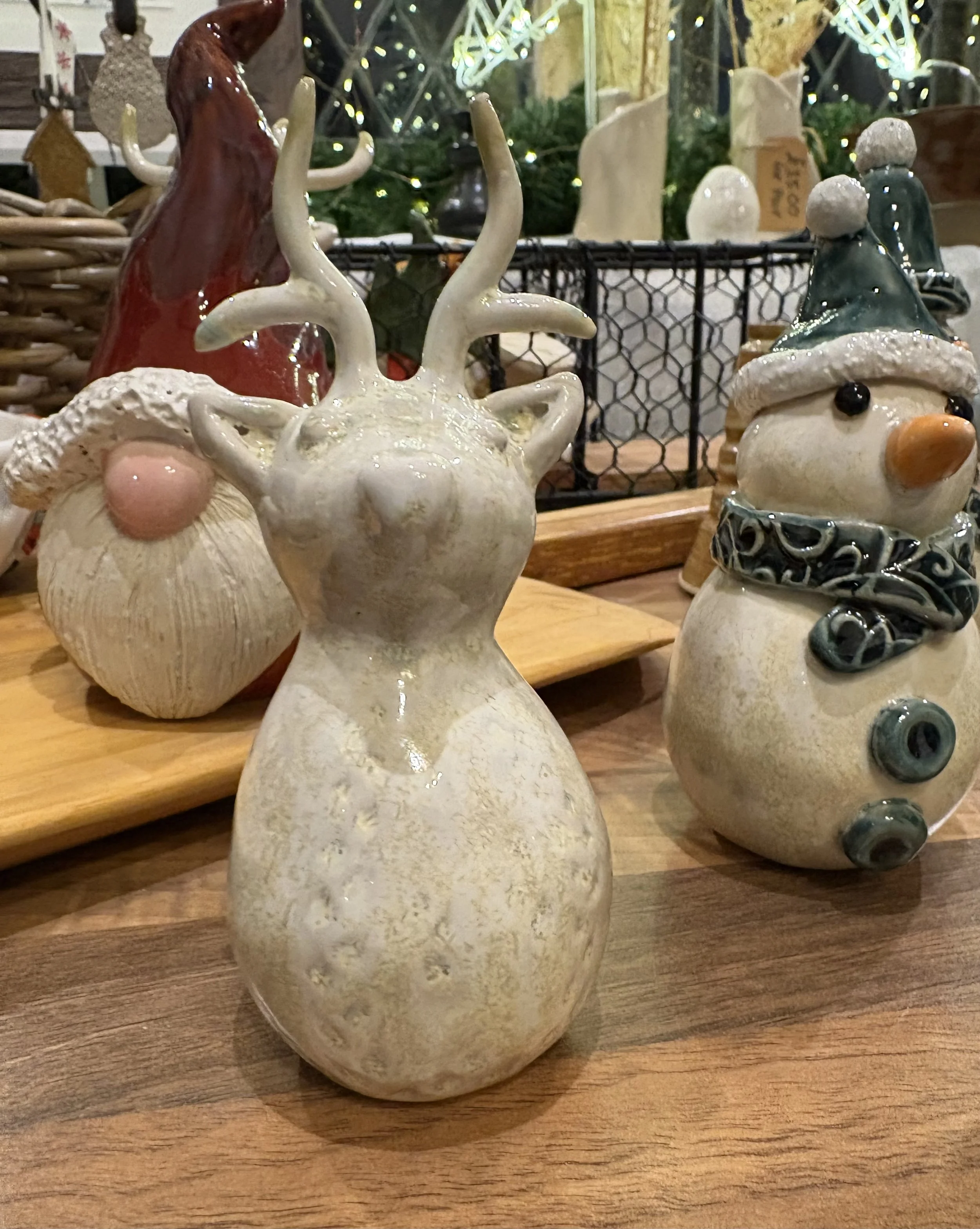 Ceramic Christmas decorations including a reindeer with antlers, a snowman wearing a hat and scarf, and a Santa face, displayed on a wooden surface with Christmas lights in the background.