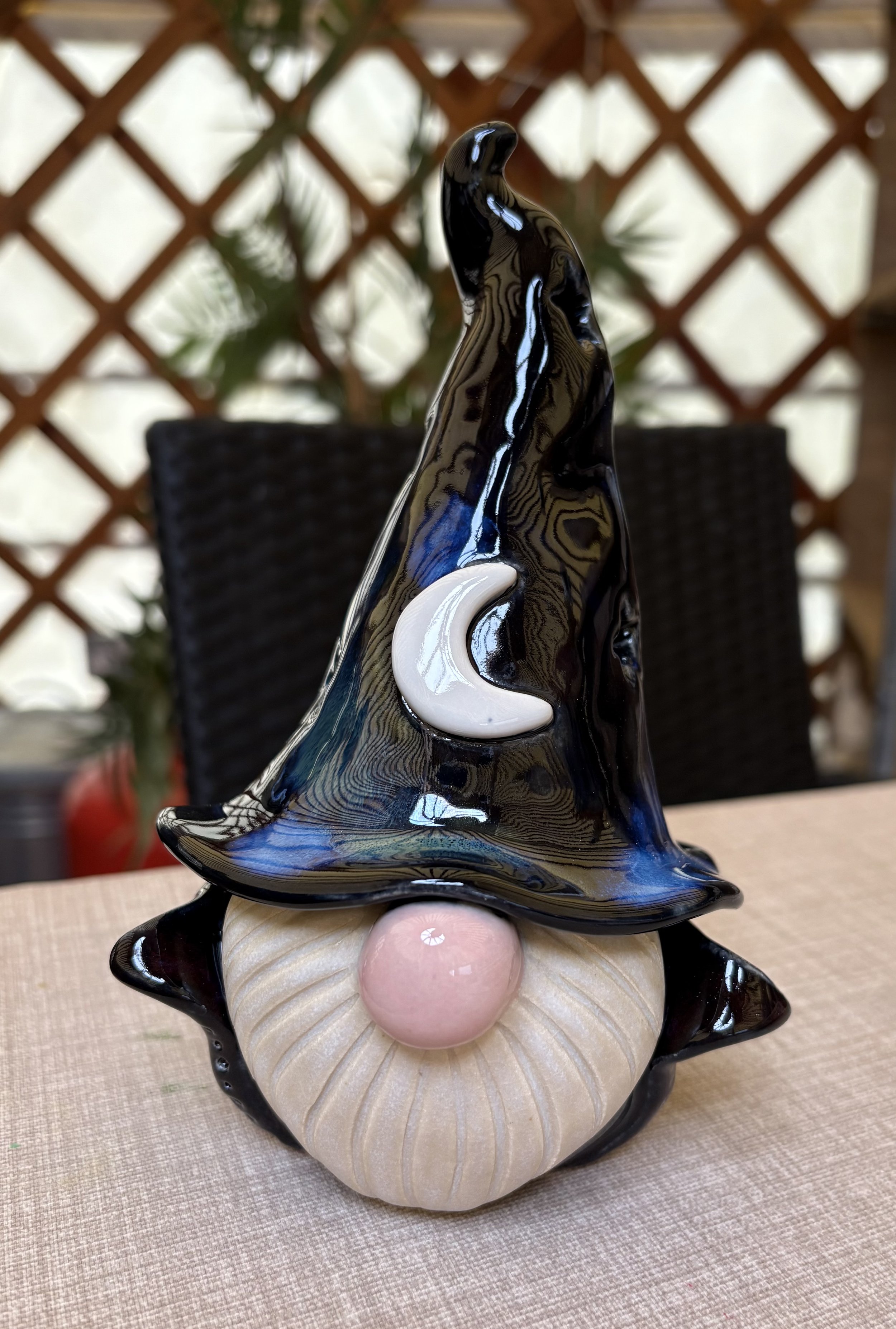 A ceramic witch sculpture with a black pointed hat, a pink nose, and a white crescent moon on its hat, sitting on a textured beige surface.