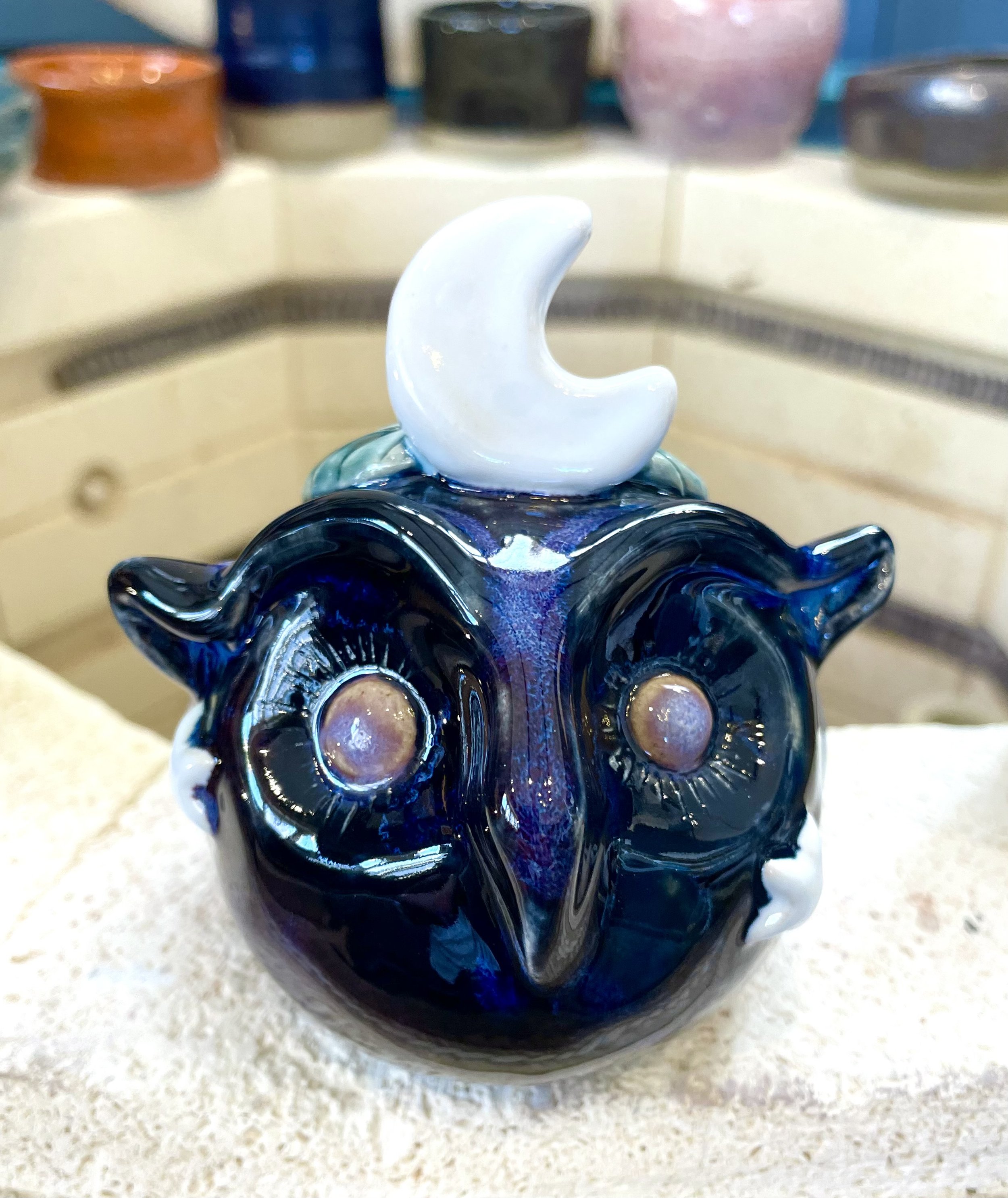 Glass art sculpture resembling a stylized owl with large circular eyes and a white crescent moon on top of its head.