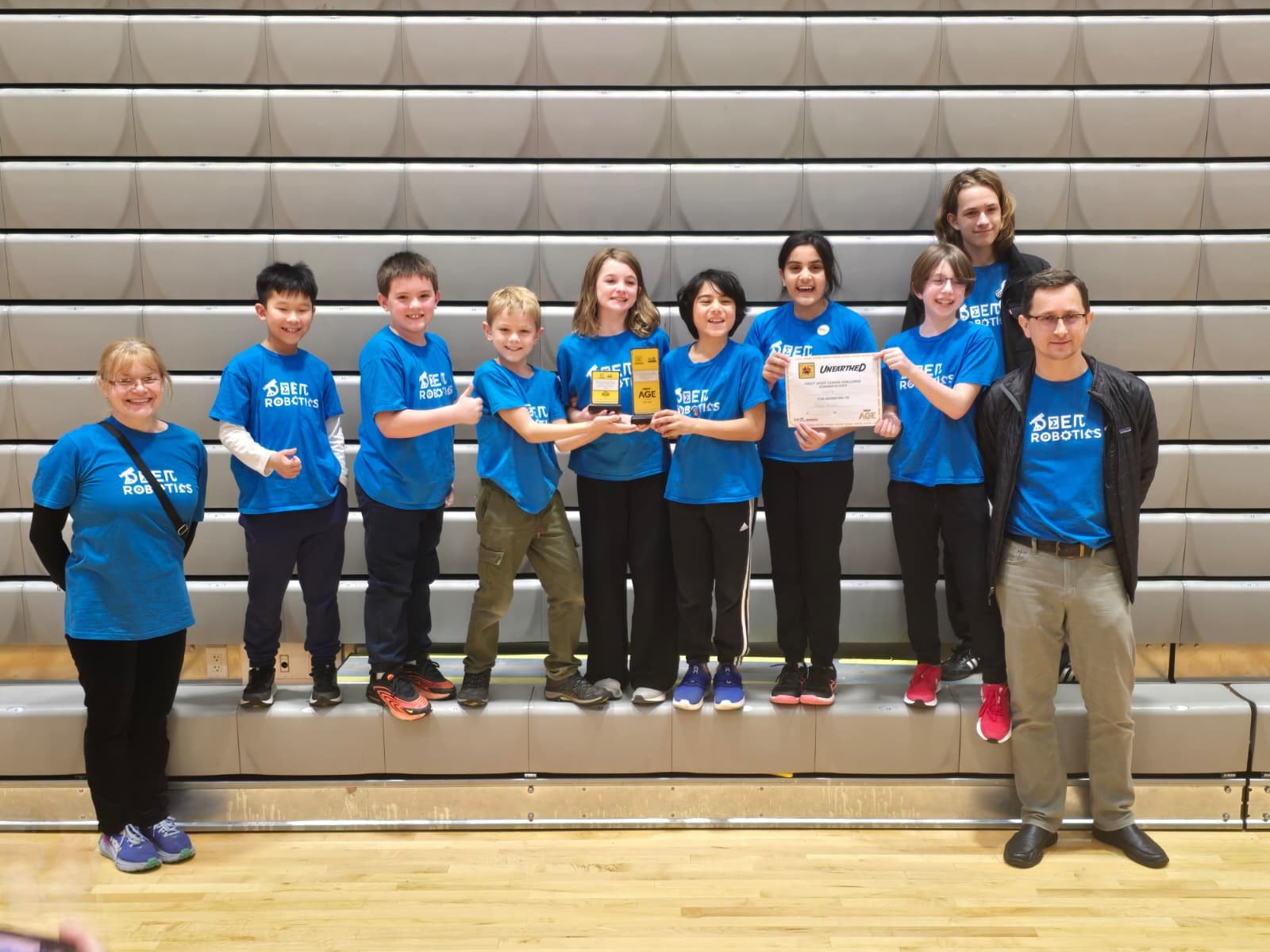 Group of young students and their coach celebrating a robotics competition win, holding trophies and a certificate, all wearing matching blue T-shirts with 'BETH ROBOTICS' logo, in an indoor gymnasium.