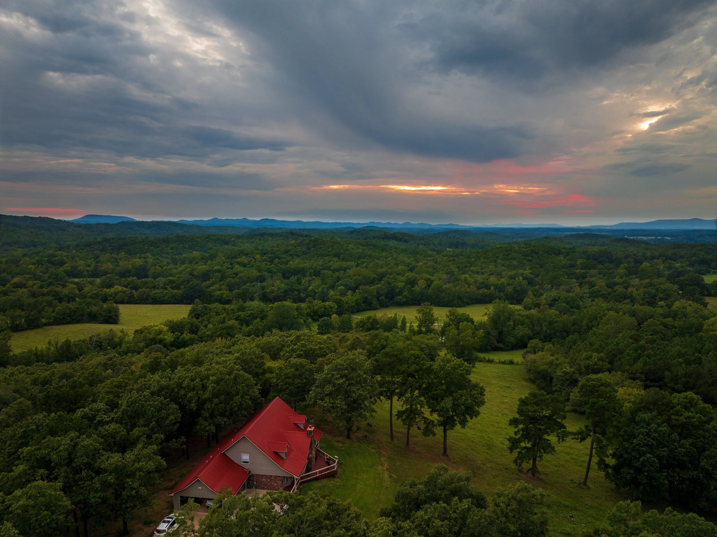 A house with a red roof surrounded by green trees in a vast landscape of green fields and dense forest, under a cloudy sky at sunset.