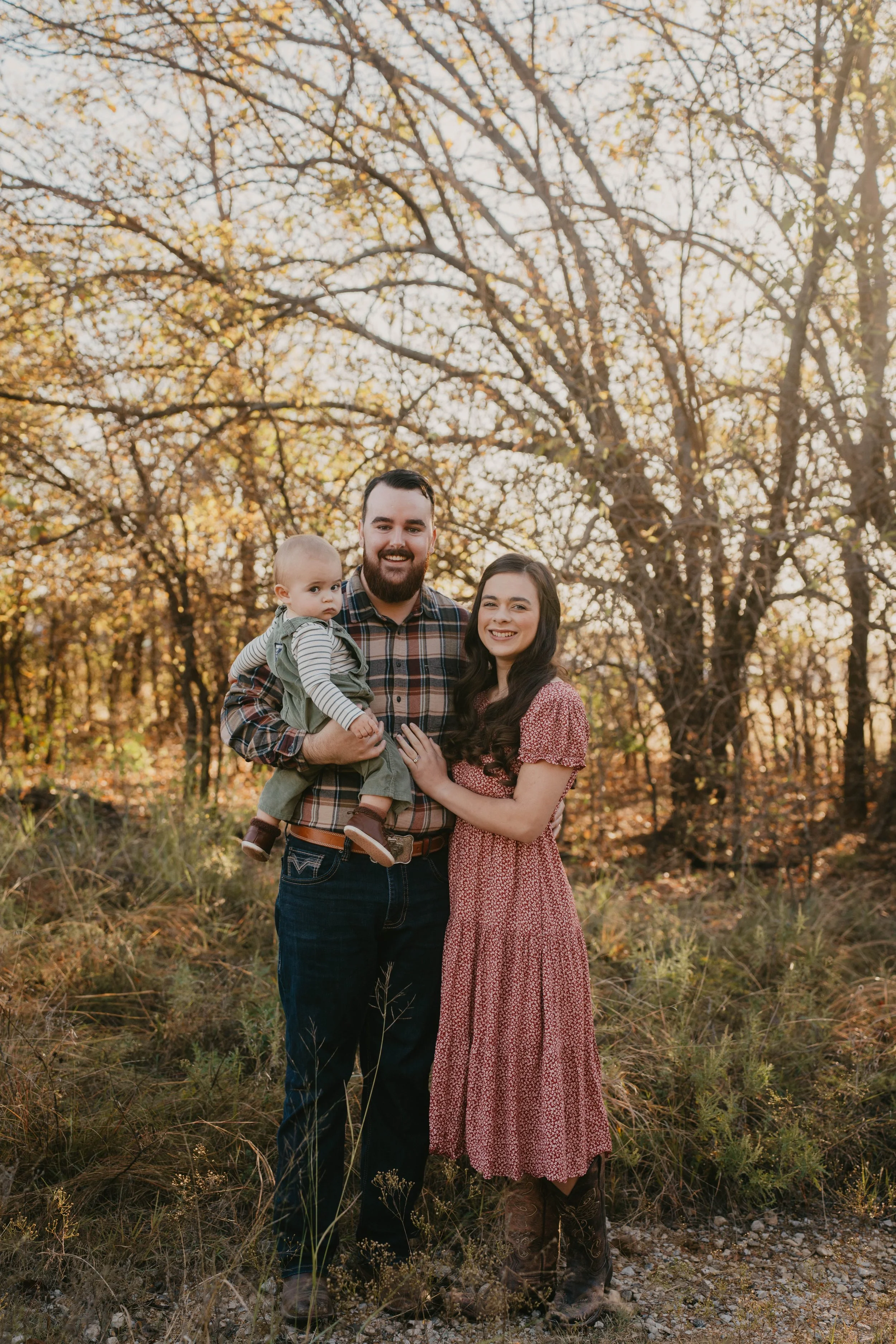 A family of three standing outdoors in a wooded area during autumn, smiling at the camera. The man has a beard and is wearing a plaid shirt, holding a young child with light hair and dressed in a striped shirt and green overalls. The woman has dark hair and is wearing a red patterned dress, standing beside the man with her hand on his chest. The background features trees with golden leaves and sunlight filtering through the branches.