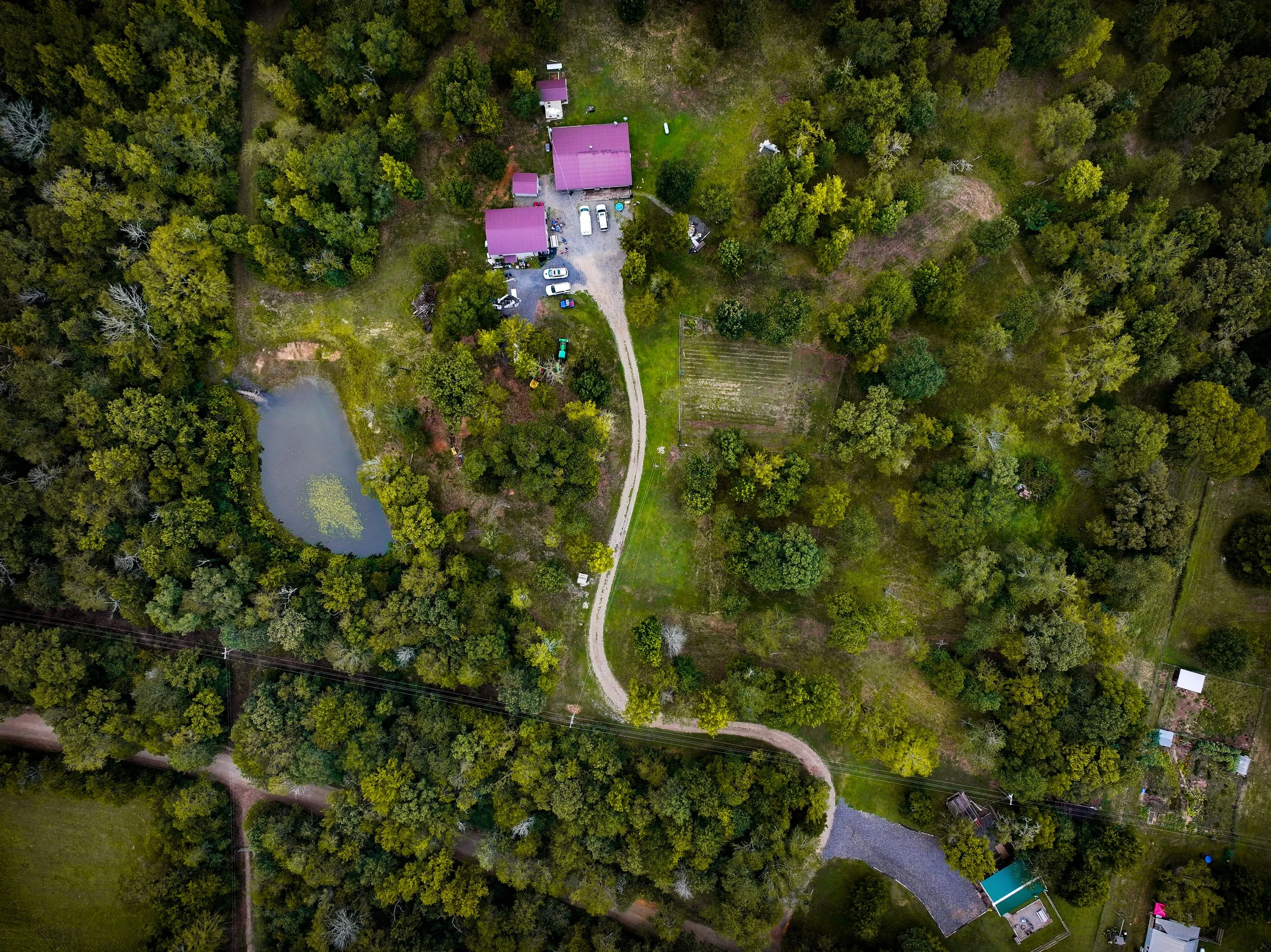 Aerial view of a rural area with a pond, a house with purple roofs, a driveway, and surrounding trees and fields.