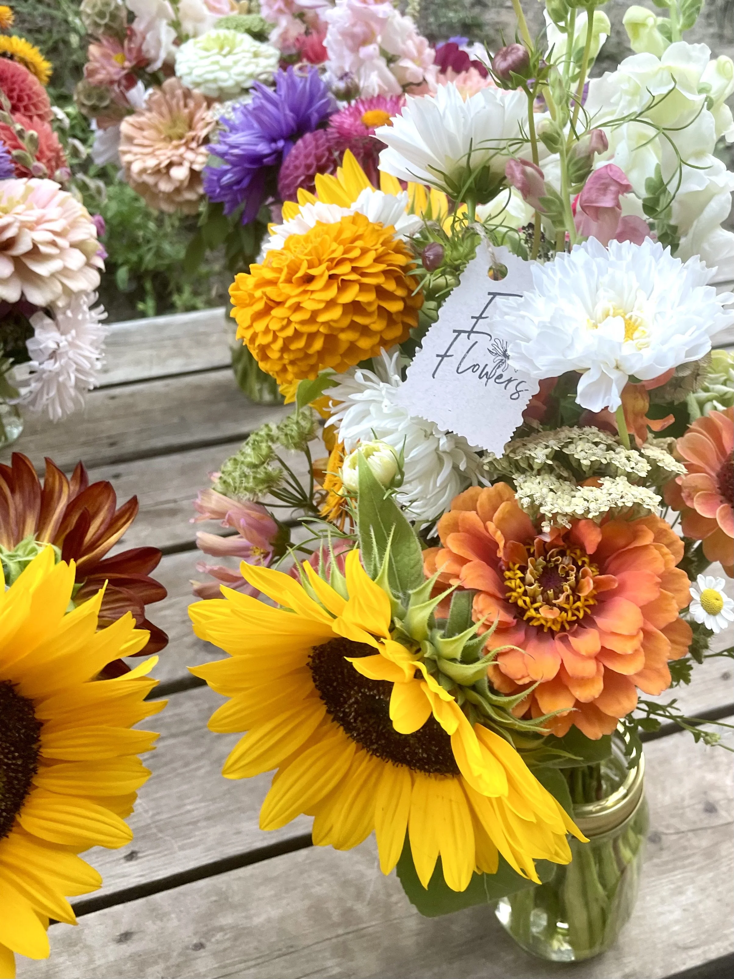 A glass jar filled with a colorful arrangement of flowers including sunflowers, marigolds, daisies, and other mixed blooms on a wooden surface.