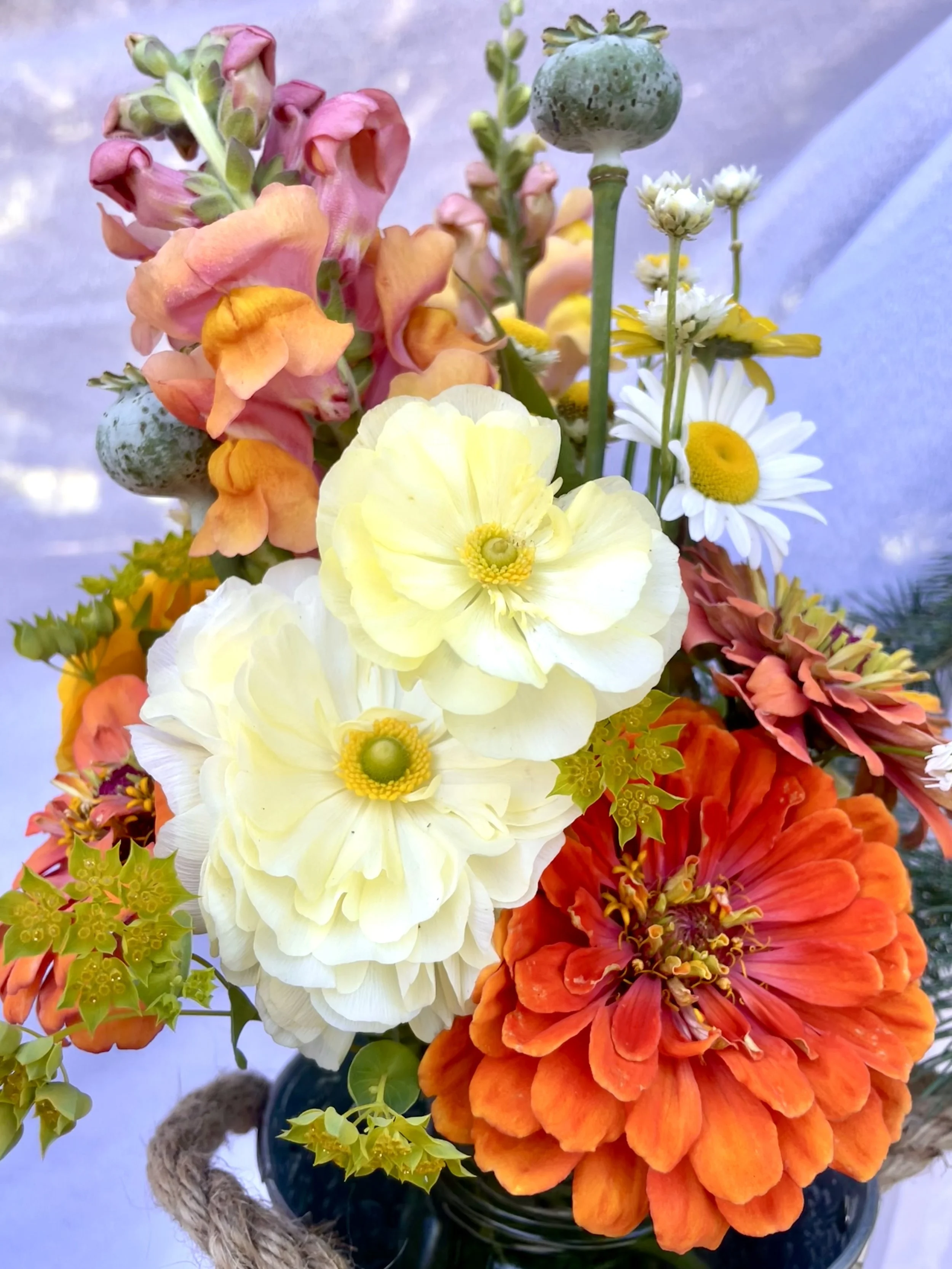 Colorful mixed floral bouquet including orange, yellow, pink, and white flowers in a dark vase with a rope handle.