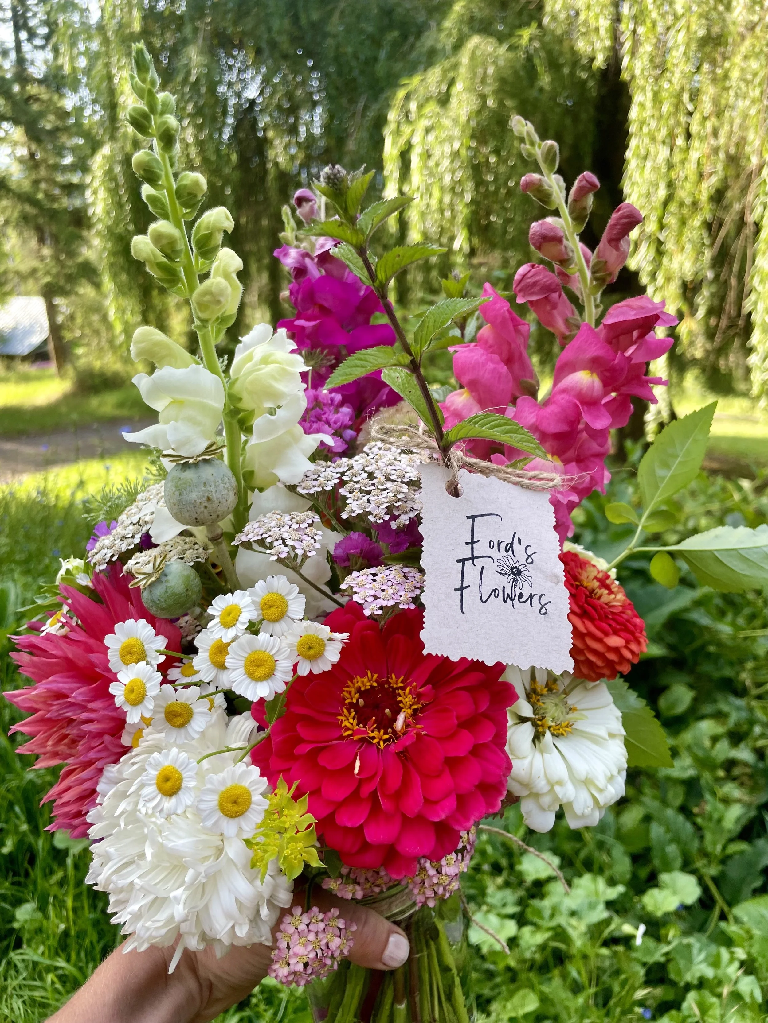 A colorful bouquet of various flowers, including daisies, dahlias, and snapdragons, with a handmade tag that reads 'Ford's Flowers', held outdoors in a garden with greenery and trees in the background.