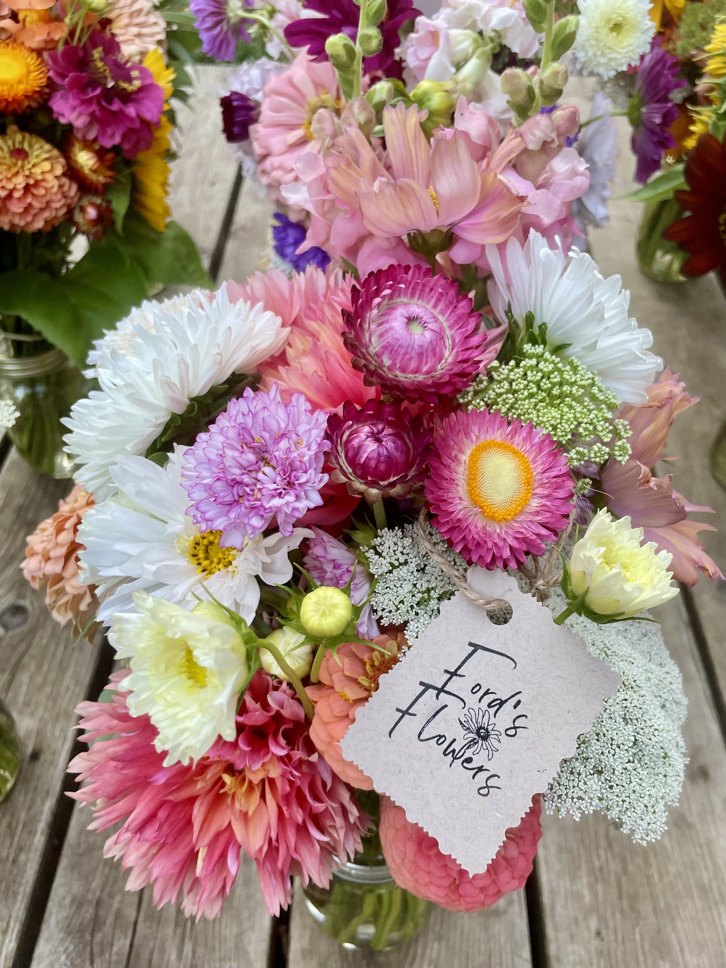 A colorful bouquet of flowers in a glass jar, including pink, white, yellow, and purple blooms, with a tag that reads 'Fordi's Flowers' attached to the arrangement.