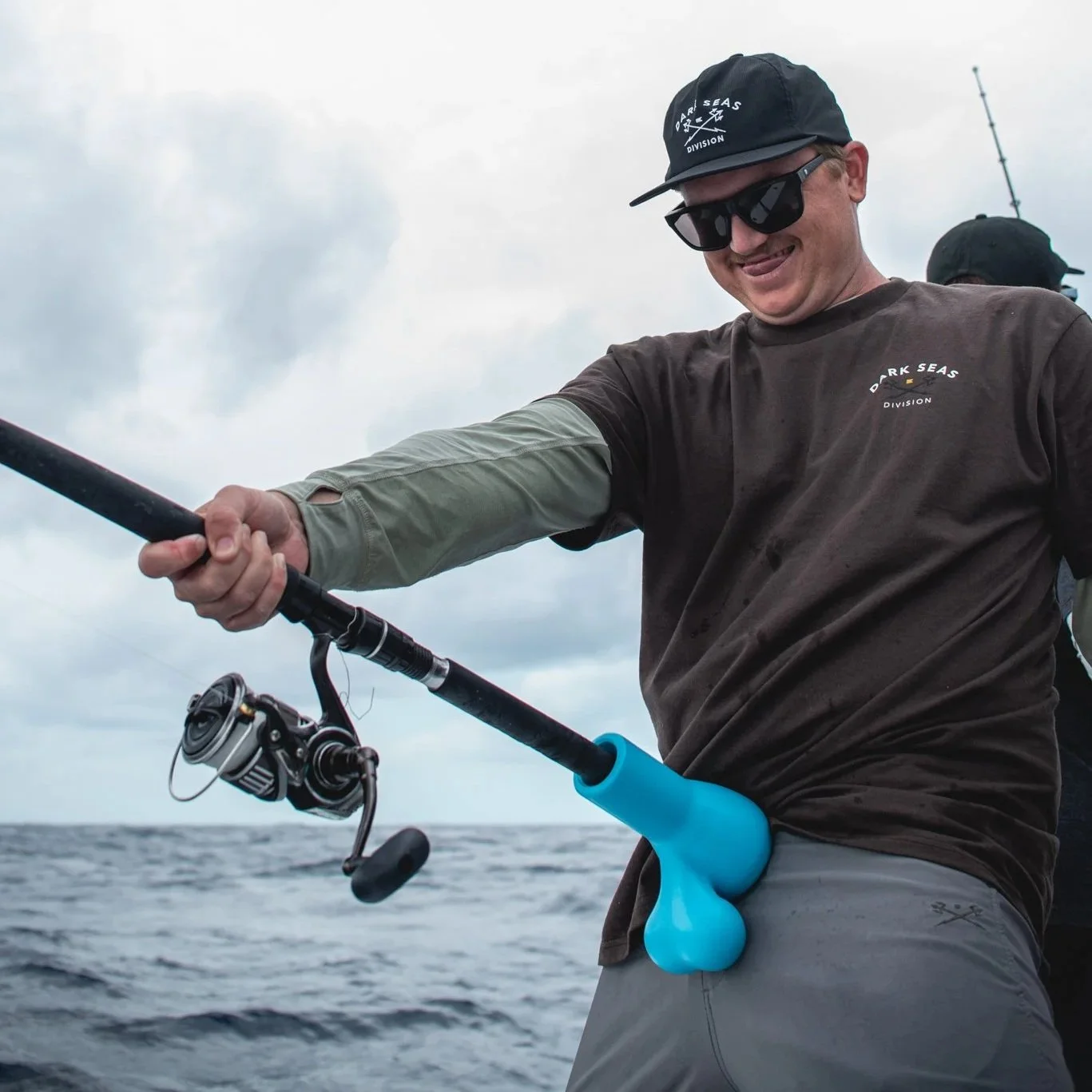 Man fishing on a boat, holding a fishing rod capped with a ball sack rod butt cushion, with cloudy sky and ocean in the background.