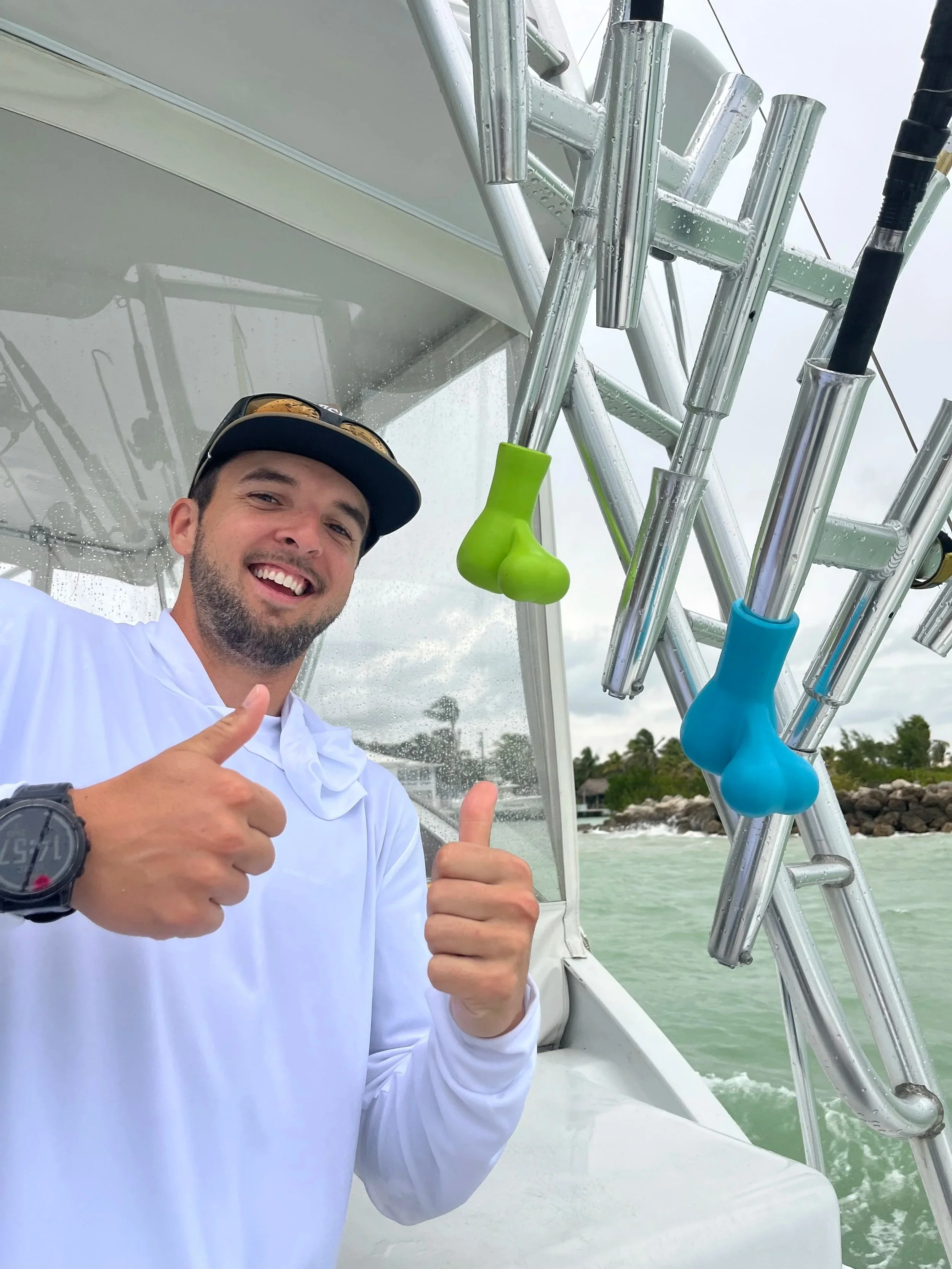 A smiling man wearing a white shirt, black cap, and a watch on his left wrist, showing a thumbs-up while standing on a boat. The boat has colorful snorkel holders and is on a body of water with a shoreline and trees in the background.