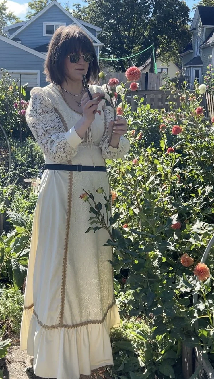 A woman with shoulder-length hair wearing sunglasses, a cream-colored, vintage-style dress with lace and embroidery, and layered necklaces, stands in a garden with various flowering plants. She is holding a flower stem and appears to be examining it.