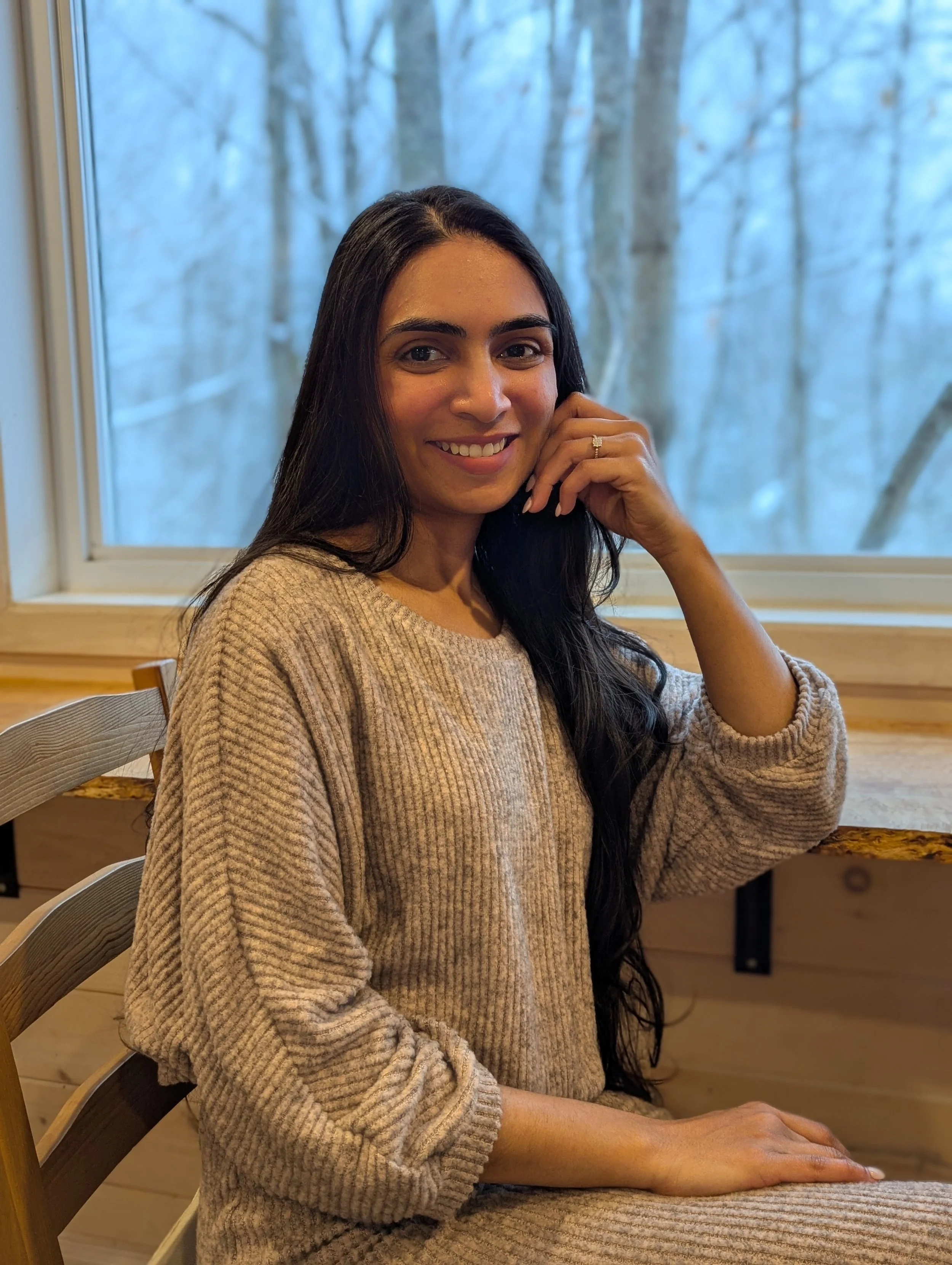 A woman with long dark hair wearing a beige knit sweater, sitting at a wooden table near a window with a snowy landscape outside, smiling at the camera.